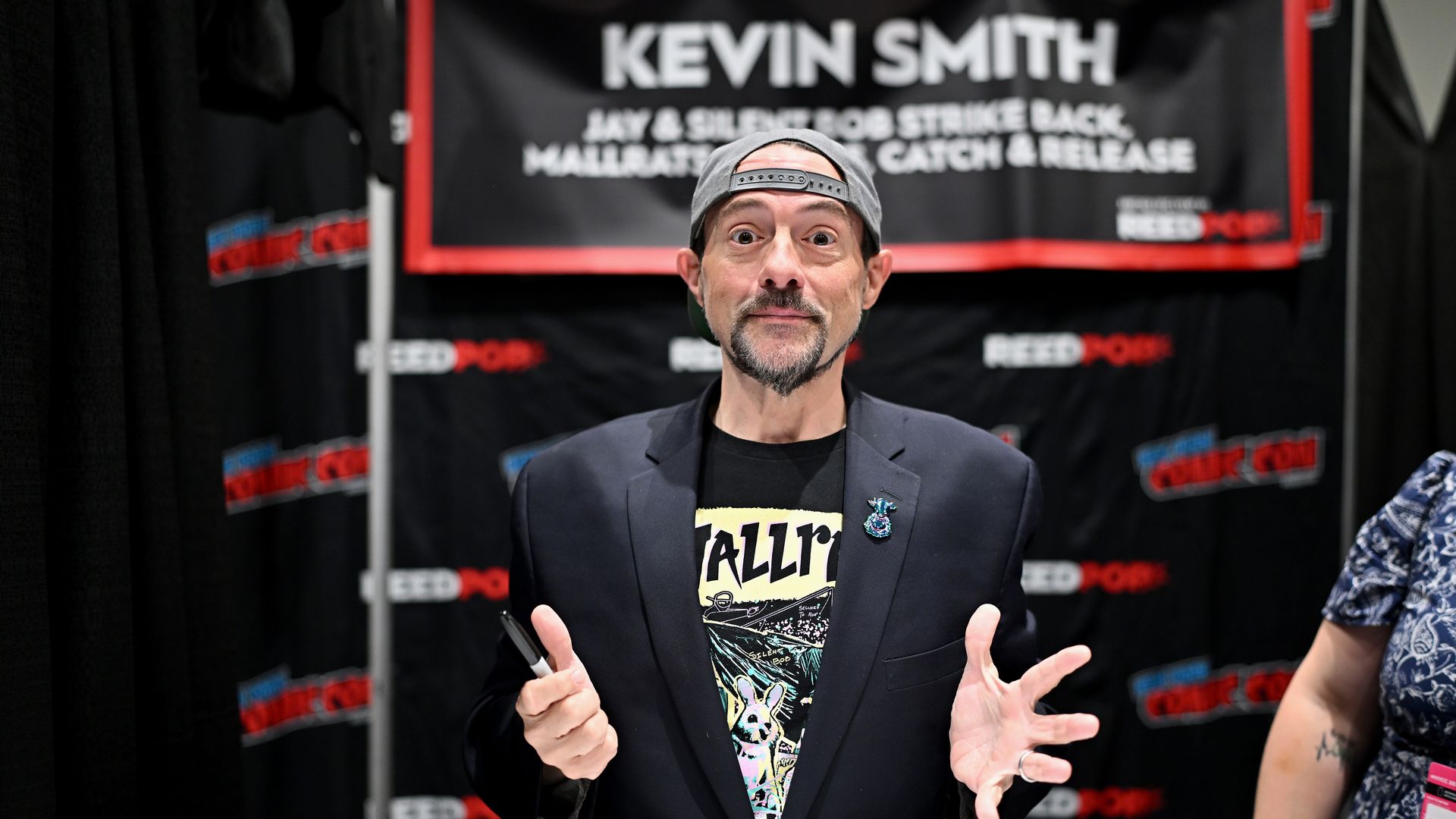 Photo of a man with a goatee posing for a photo with the name "Kevin Smith" on a sign above him. 