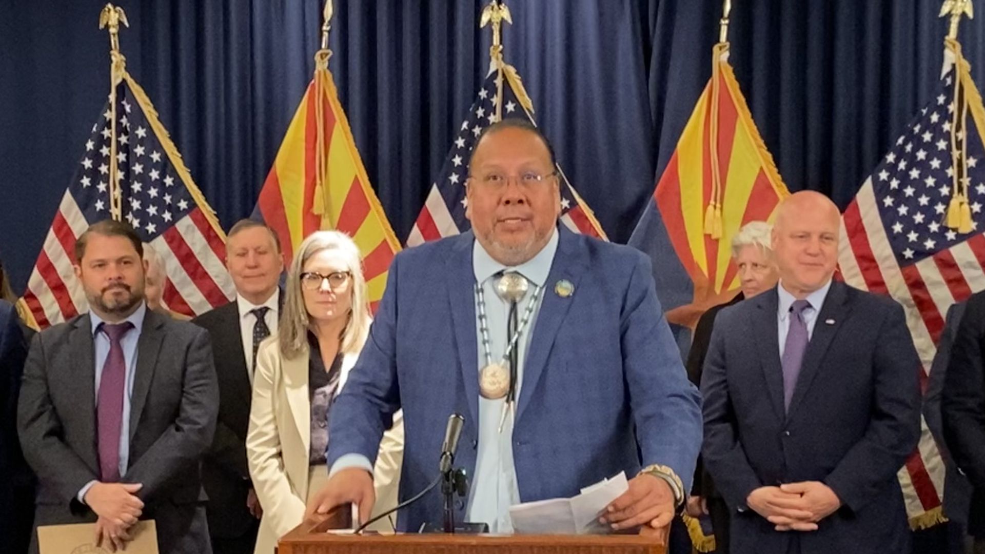 A man in a sport coat and a bolo tie speaks at a podium with several people and a row of US and Arizona flags behind him. 
