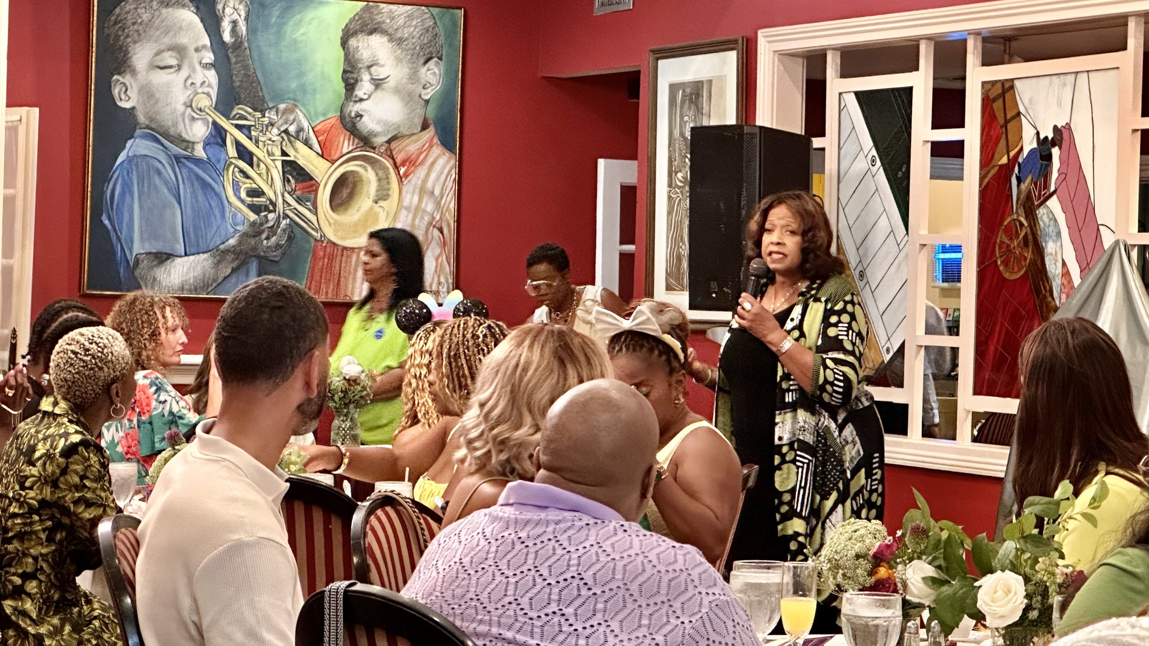Woman holding a microphone speaking to a seated audience in a red-walled room with a large painting of children playing trumpets, and floral centerpieces on tables.