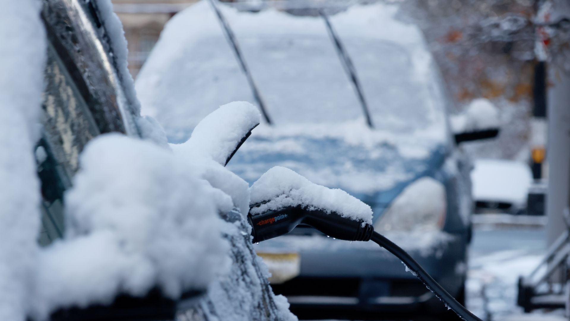 A close-up of a snow-covered electric vehicle, plugged into a charger, with the charging cable also covered in snow. 
