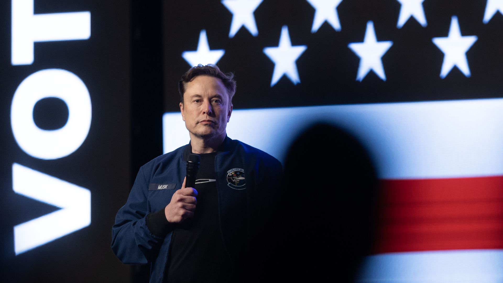 Elon Musk holding a microphone in front of an American flag.