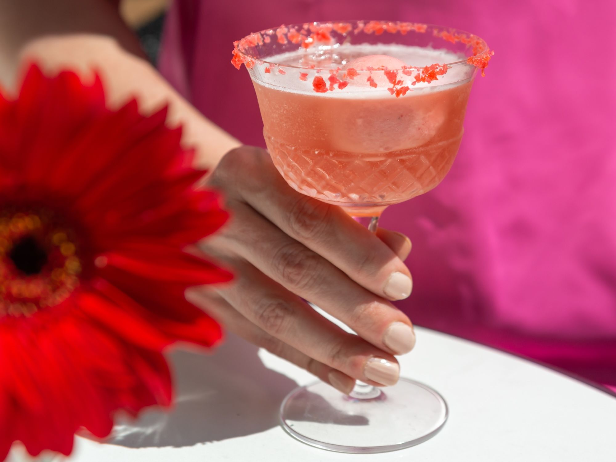 Hand with nude nail polish holding a pink cocktail in a glass with red salt rim, next to a bright red flower on a white surface, bright pink background.