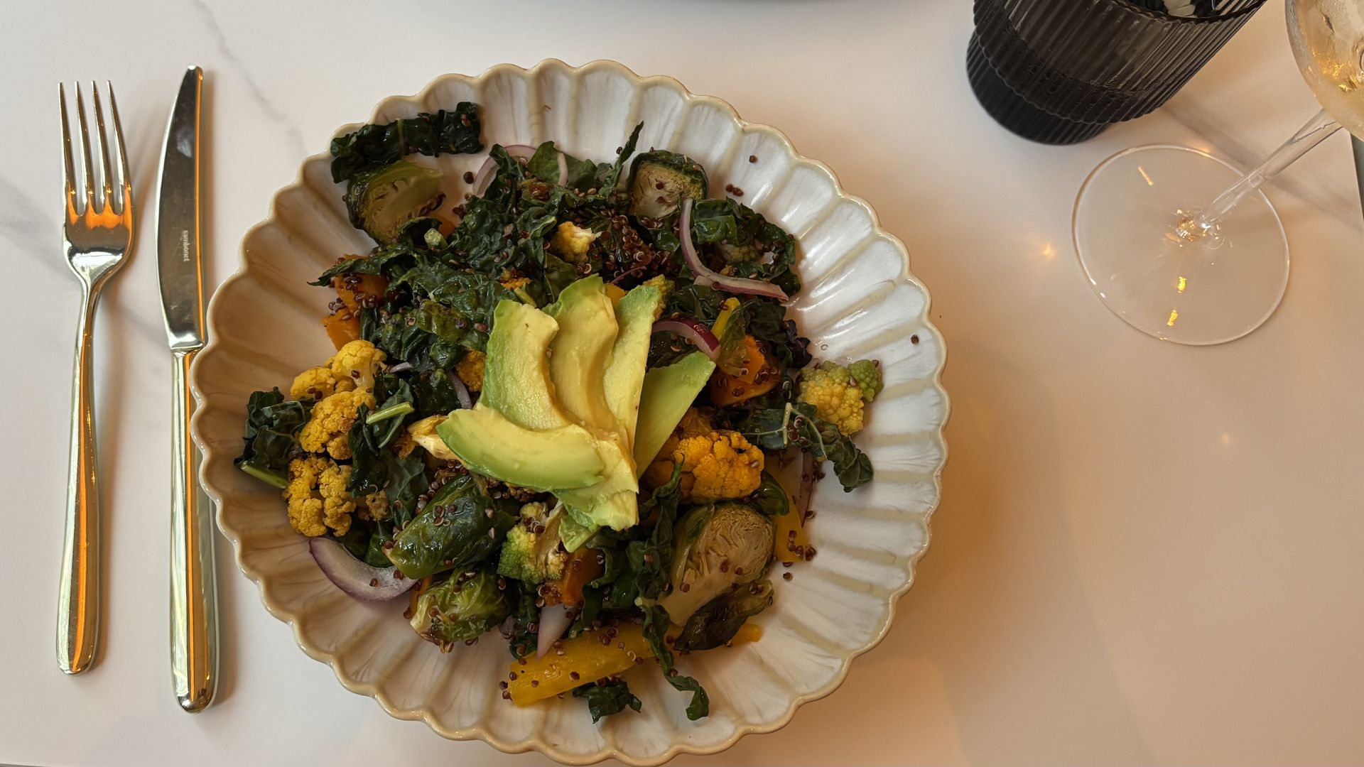 Plate of mixed salad with kale, avocado slices, roasted cauliflower, Brussels sprouts, red onions, and quinoa, placed on a white scalloped plate with a knife and fork nearby on a white table.