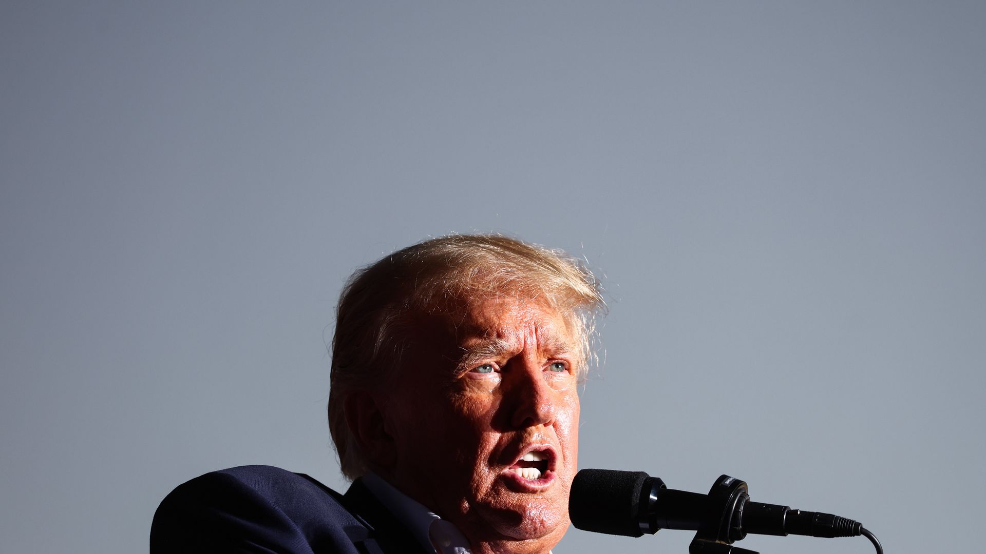 Donald Trump, wearing a blue suit and white shirt, speaks into a microphone at a campaign rally underneath an overcast sky.