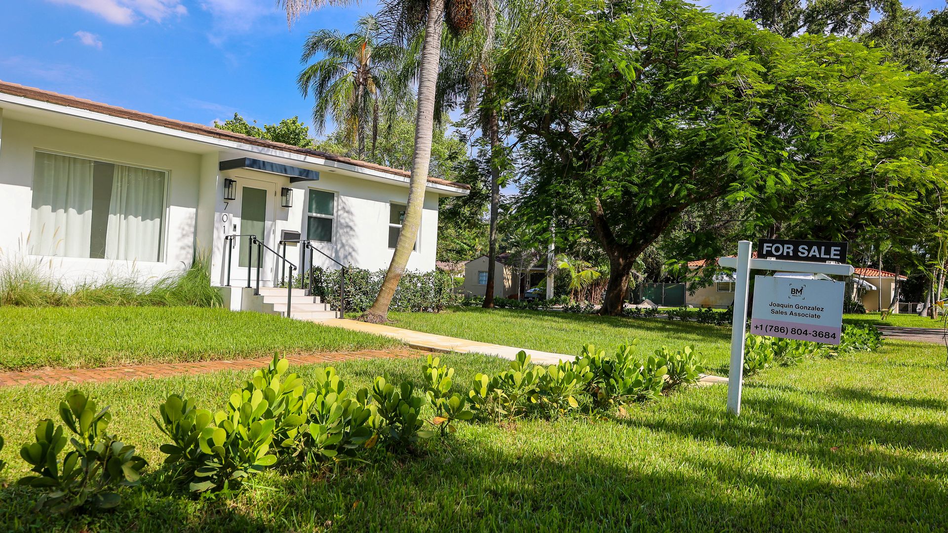 Single-story white house with a tiled roof, palm tree, green lawn, and a "For Sale" sign listing Joaquin Gonzalez with a phone number under a clear blue sky.