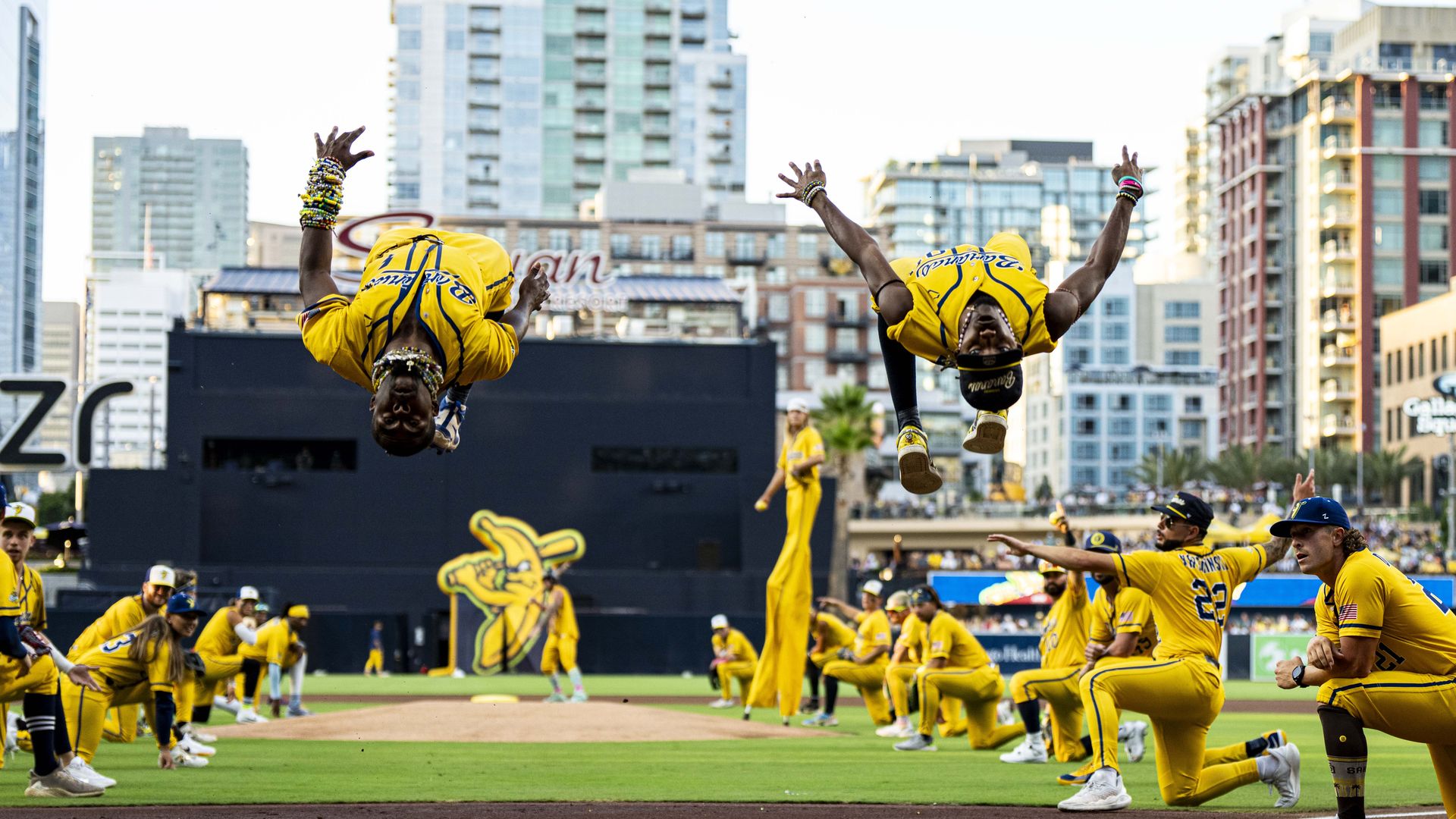 Two players in bright yellow baseball uniforms flip midair with arms raised on a grassy field, as teammates cheer nearby; a city skyline looms in the background.