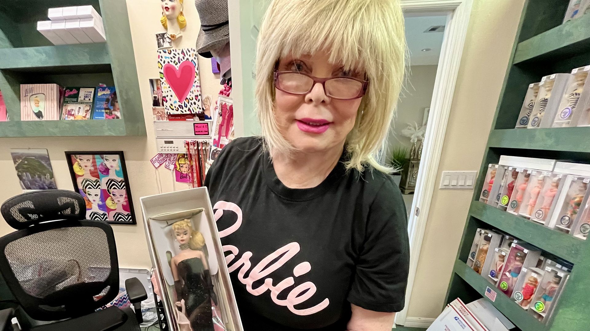 A blonde woman in a black T-shirt that says "Barbie" in pink across the front holding a blonde Barbie doll in a black mermaid gown.