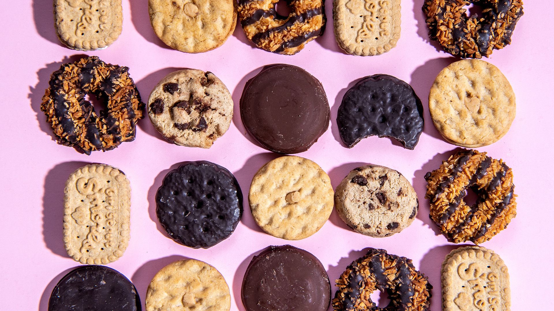 Assorted Girl Scout cookies on a pink background, including Samoas with caramel and chocolate, Thin Mints, Tagalongs, Trefoils, and chocolate chip cookies arranged in a grid pattern.