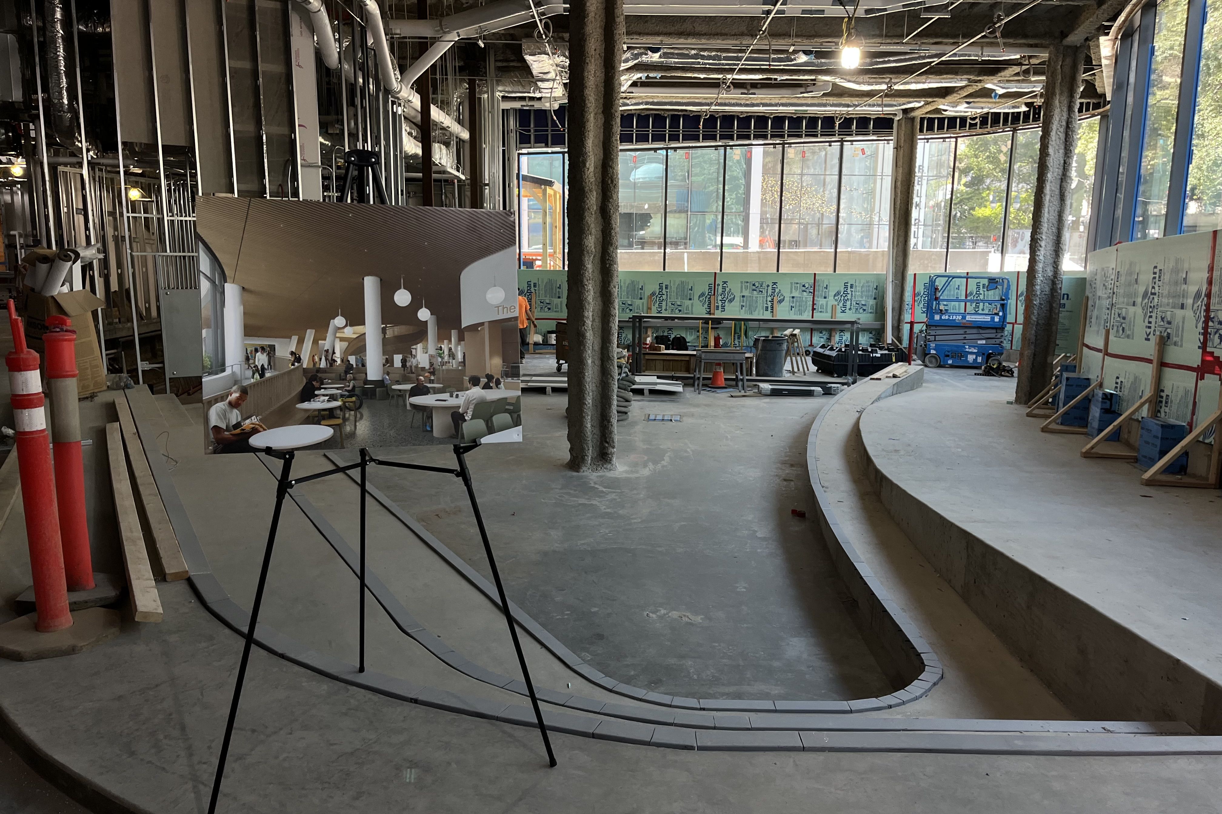 Indoor construction site with exposed ceiling and insulation, a curved seating area, and a rendering of finished space on a stand showing people sitting and round tables under pendant lights.