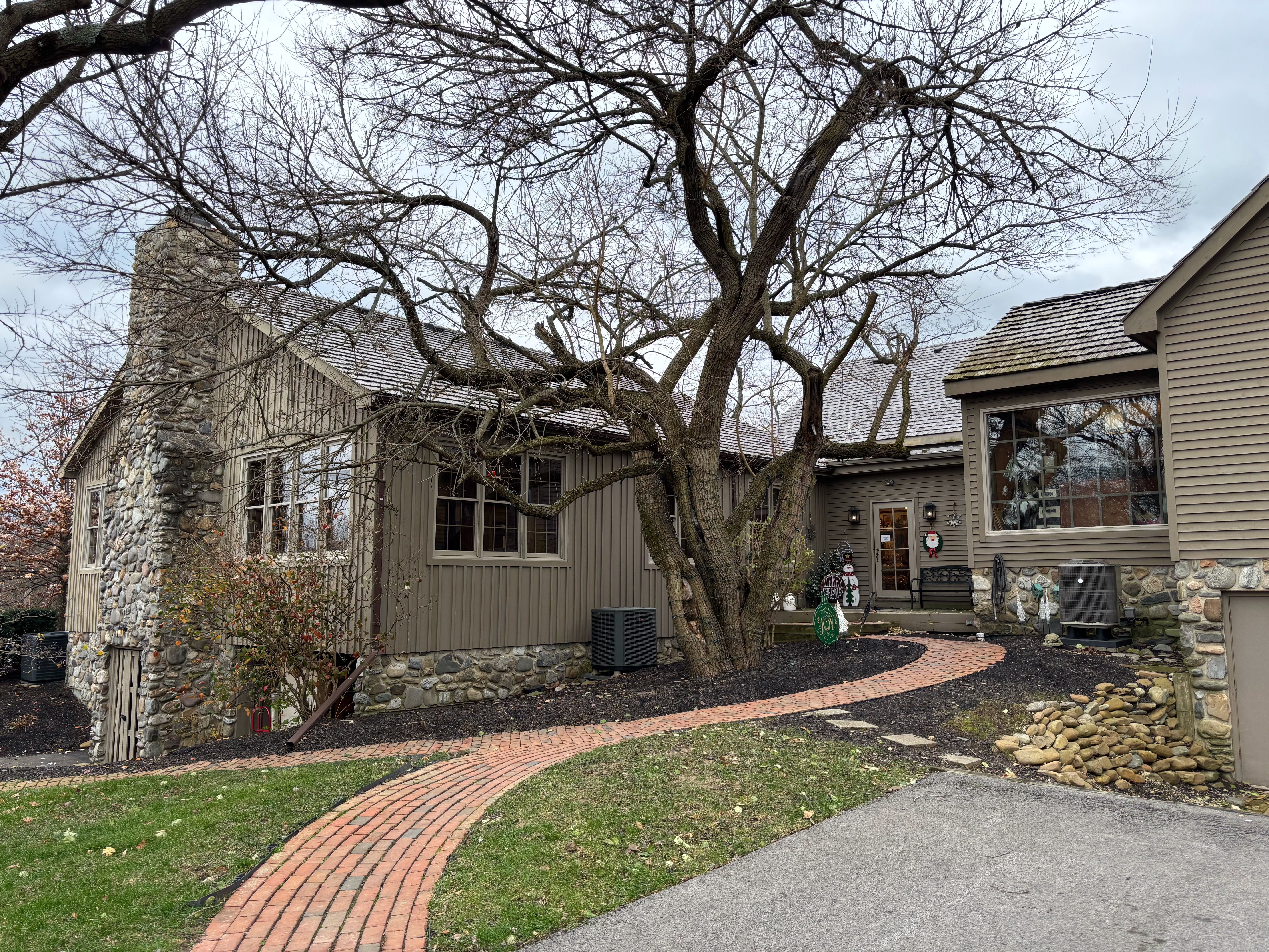 Grayish-brown house with stone chimney and foundation, leafless tree in front, curved brick walkway, and Christmas decorations on the door and near entrance on a cloudy day.