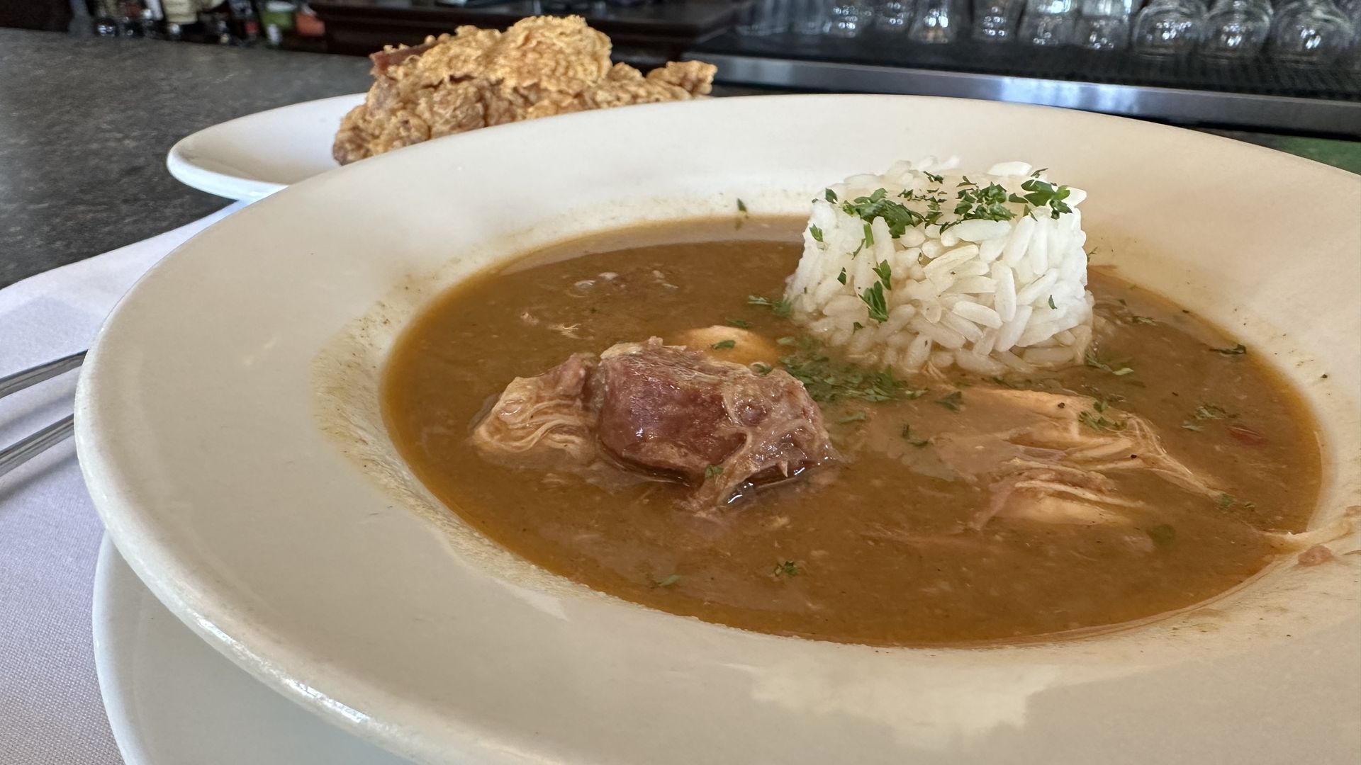 A bowl of gumbo is seen in the foreground, with strips of chicken and sausage visible in the broth, and rice in a cylinder-shape off to the right side. In the background, a fried chicken thigh can be seen on a plate.