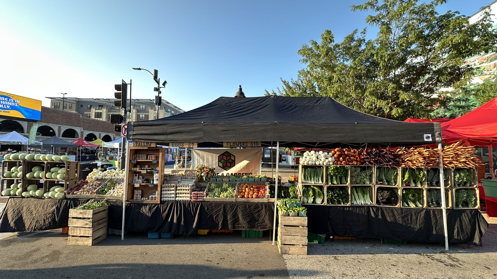 Outdoor farmers market stalls under a large black canopy on a sunny day. Crates and tables display watermelons, onions, and vegetables, with a blue sky and leafy trees in the background.