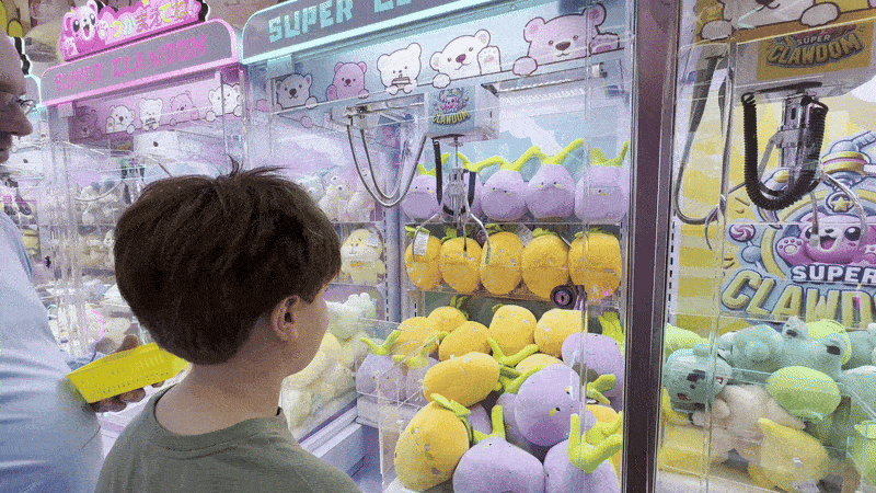 A claw machine picks up a stuffed carrot and drops it while a boy and his dad react.