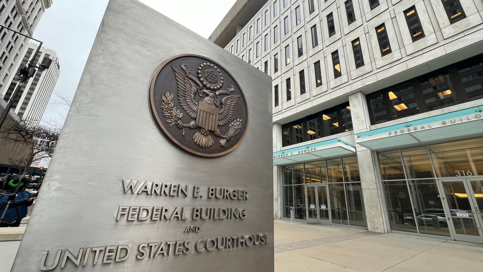 Bronze U.S. seal with eagle on a gray monument in the foreground; engraved title reads "Warren E. Burger Federal Building and United States Courthouse." A modern, multi-window federal building dominates the background.