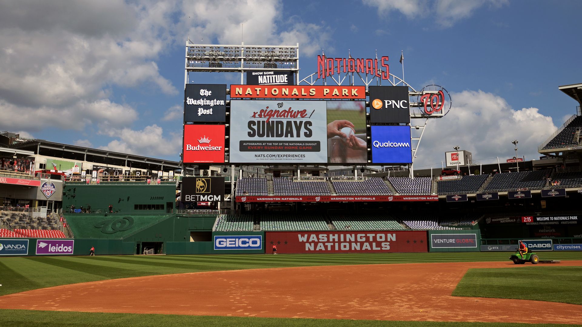 Wide view of Nationals Park with a large scoreboard, sponsor boards, and banners. Blue sky with clouds above the field; dirt infield and green outfield, Washington Nationals signage along the wall.