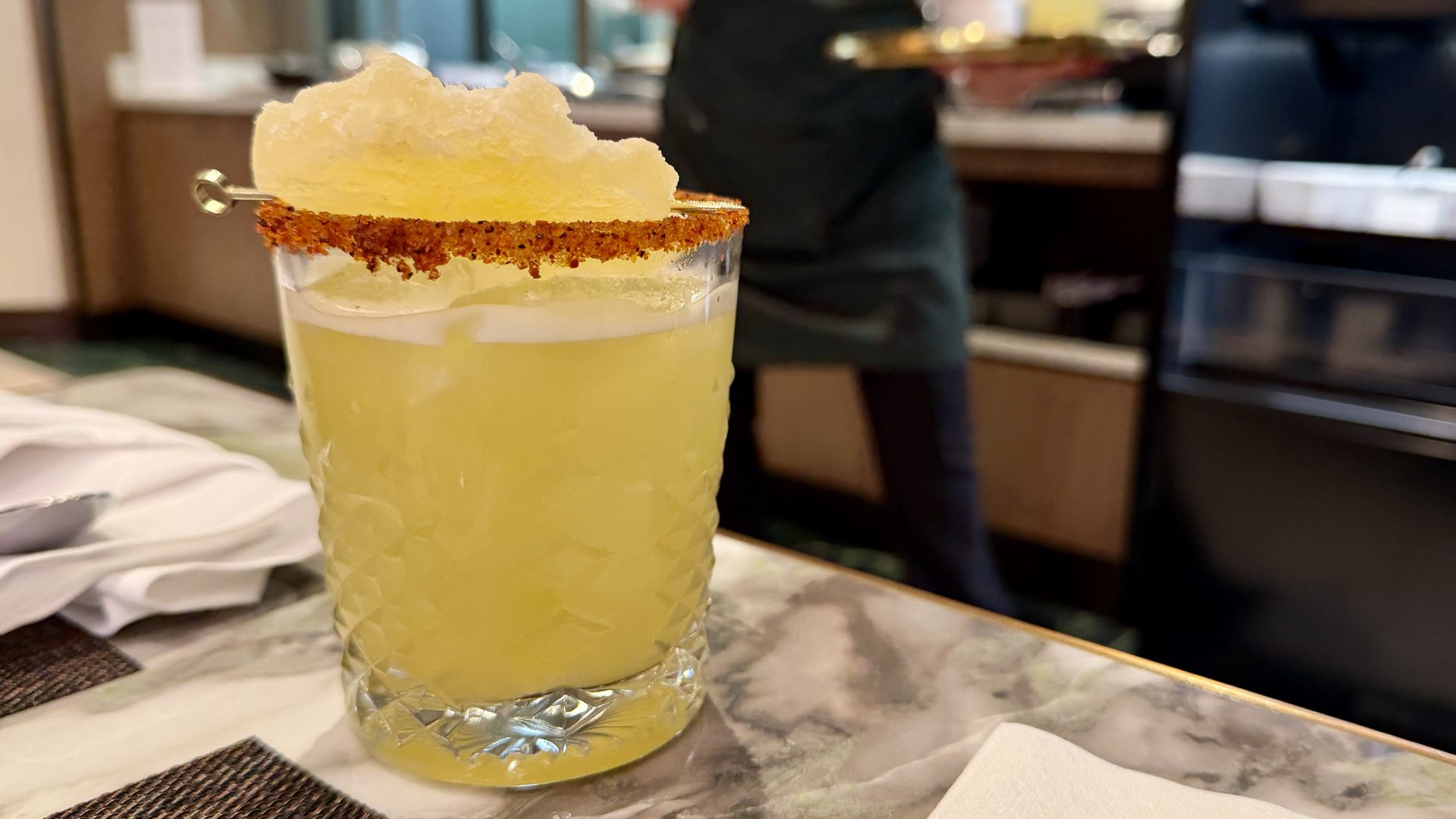 Pale yellow cocktail in a textured glass with a brown sugared rim and a large white cotton-candy garnish, set on a marble table in a busy restaurant.