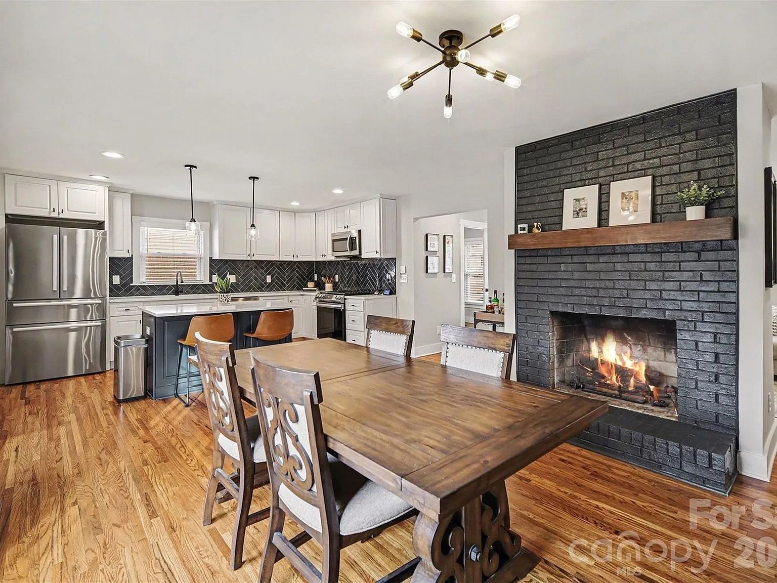 Modern kitchen and dining area with wooden floors, a wooden dining table with six chairs, a black tiled fireplace with fire, white cabinets, stainless steel appliances, and pendant lights over a kitchen island.