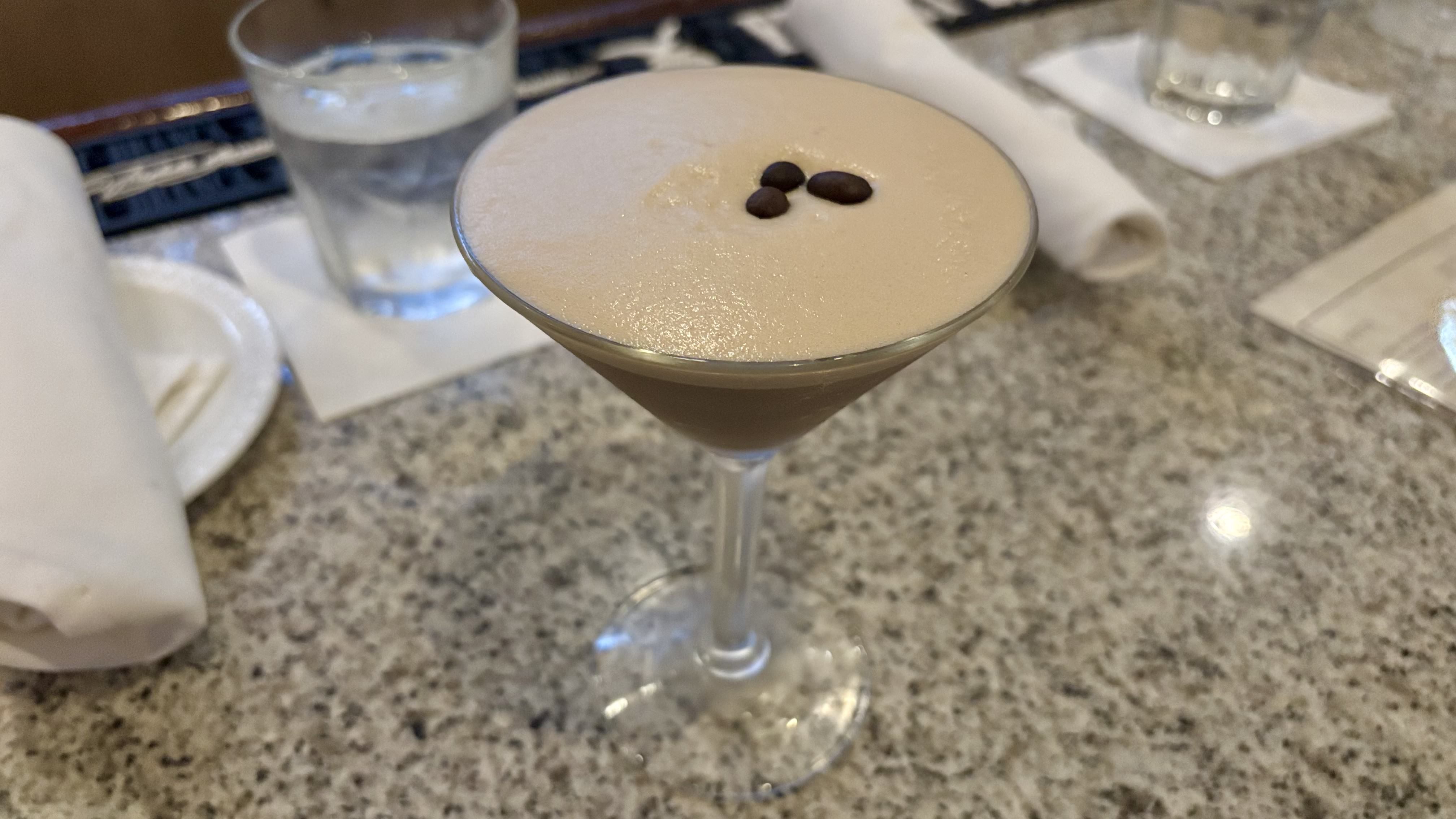 Creamy espresso martini in a clear martini glass topped with three coffee beans, placed on a speckled countertop with glass of water and white napkins in the background.
