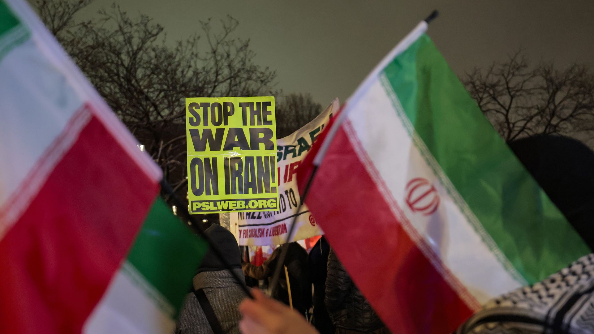 A lime green sign that reads in all-caps black letters "STOP THE WAR IN IRAN" can be seen in a crowd of protesters with Iranian flags.