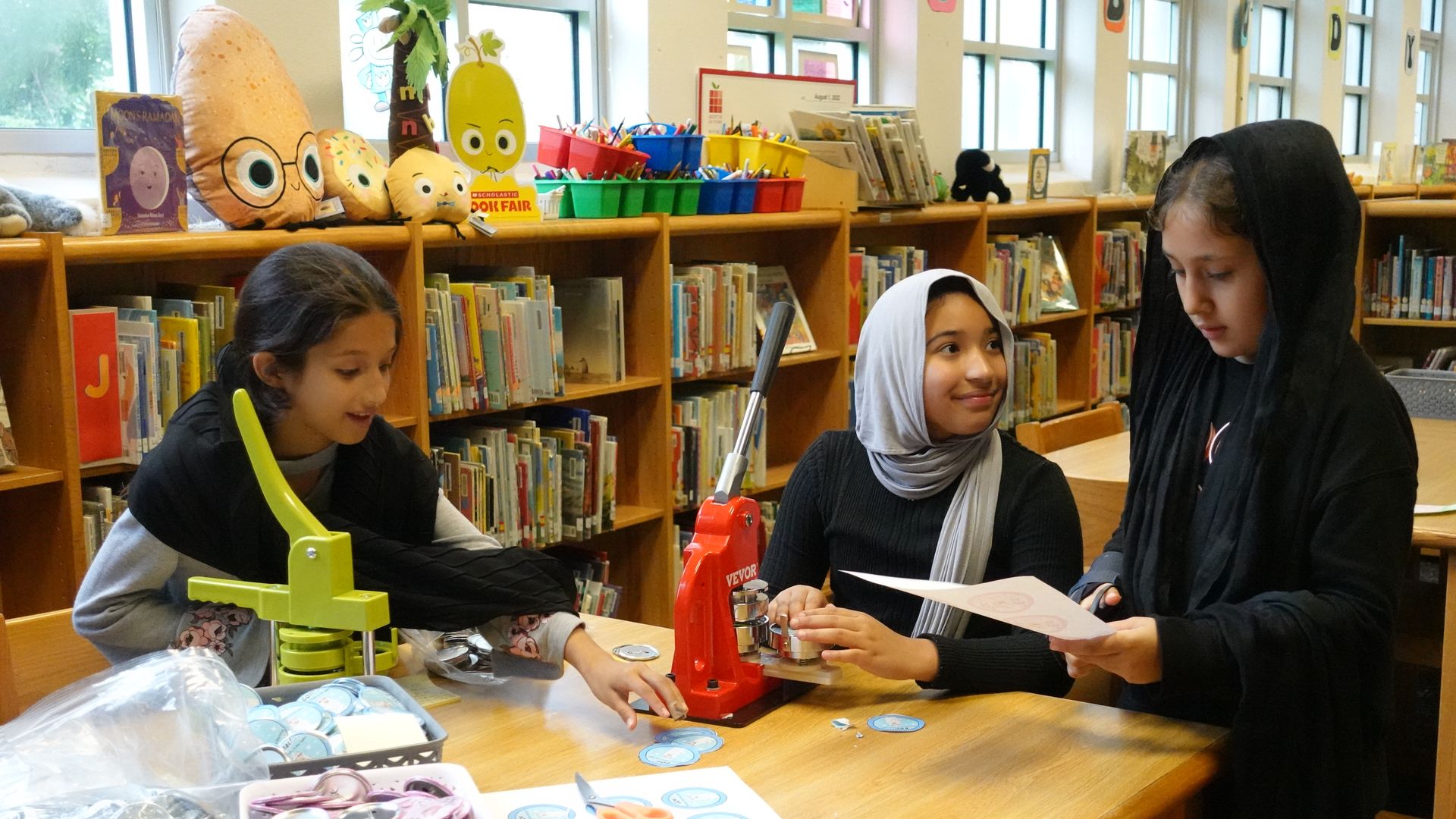 A photo of the three girls designing their pins with library books in the background.