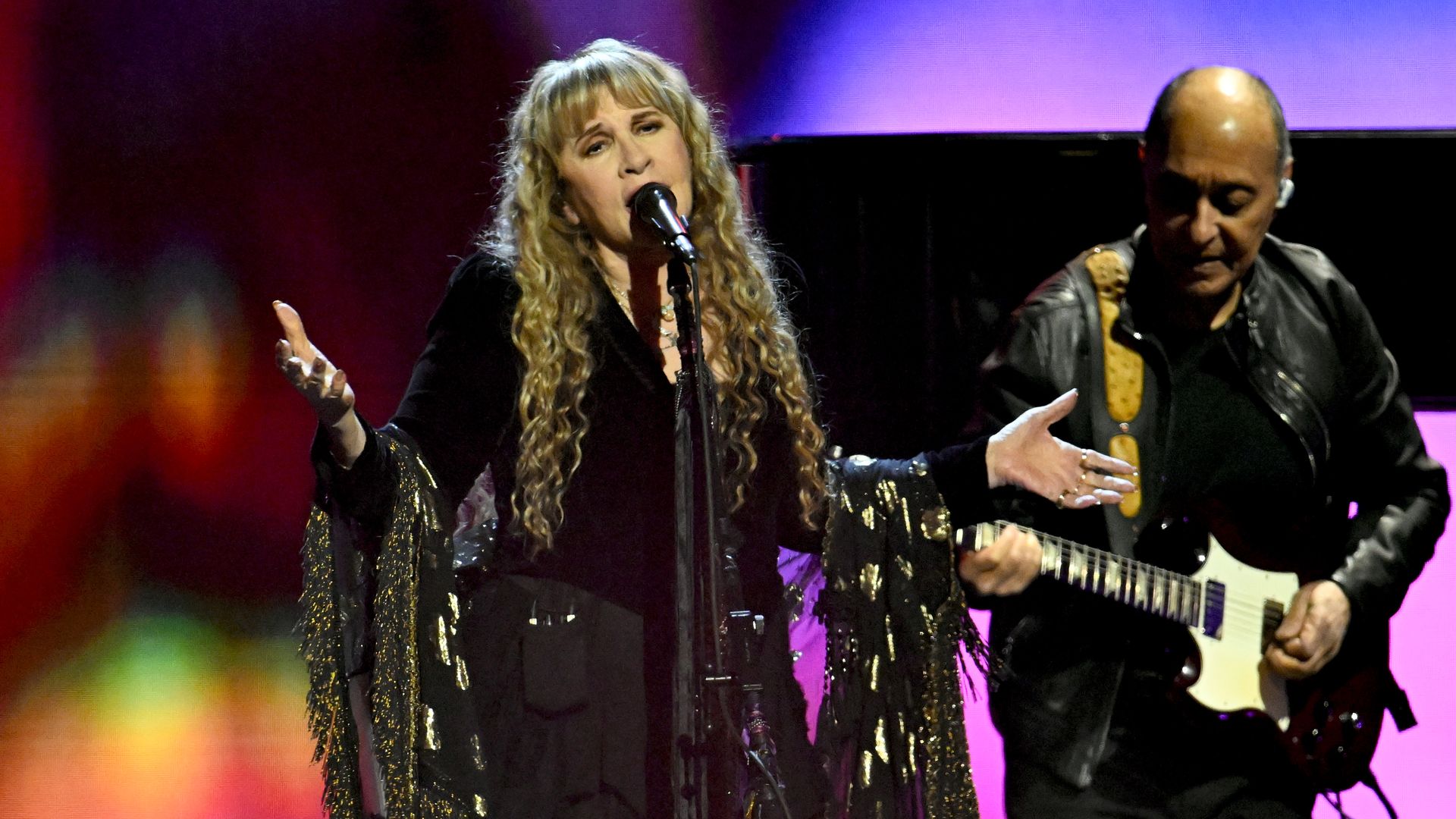 A woman with long, curly blonde hair wearing a black top with flowy bell sleeves sings into a microphone.