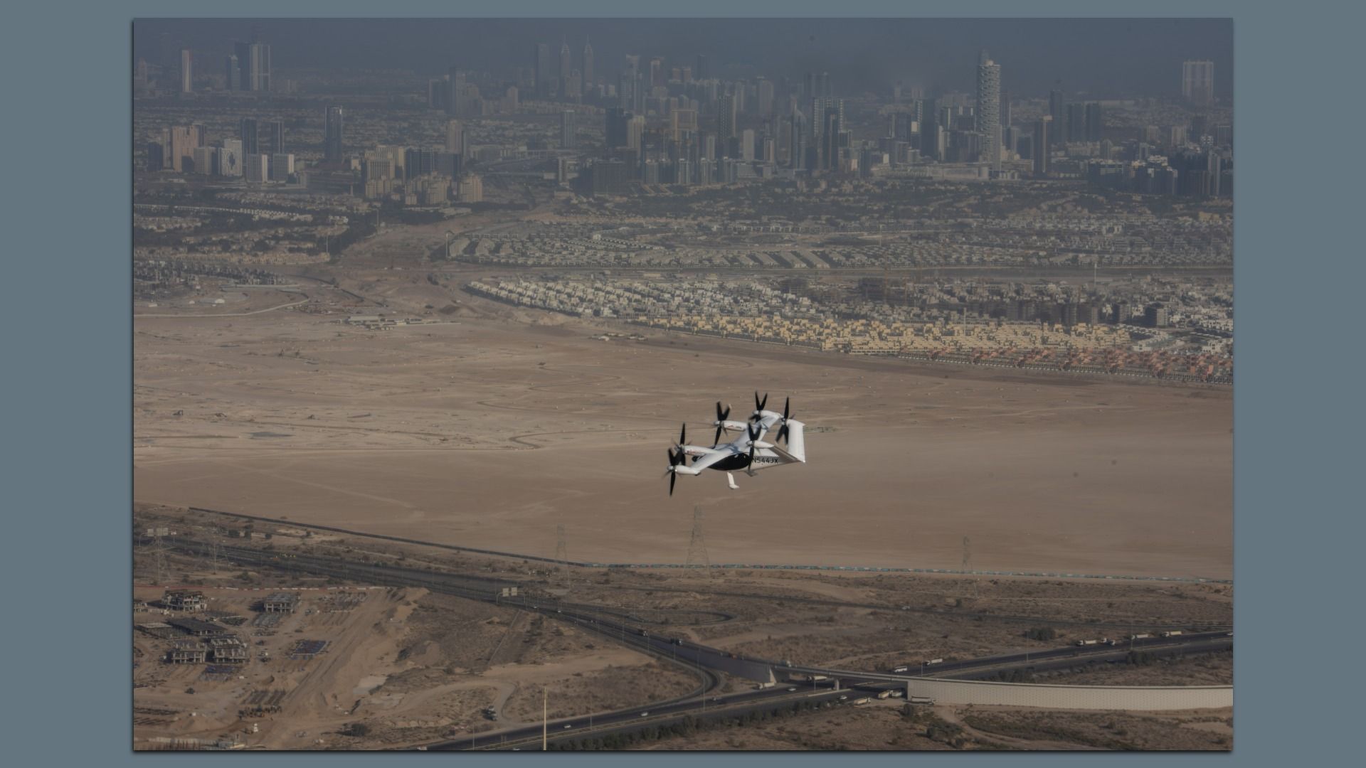 White and black electric vertical takeoff aircraft flying over a desert area with roads and cityscape in the background under hazy sky.