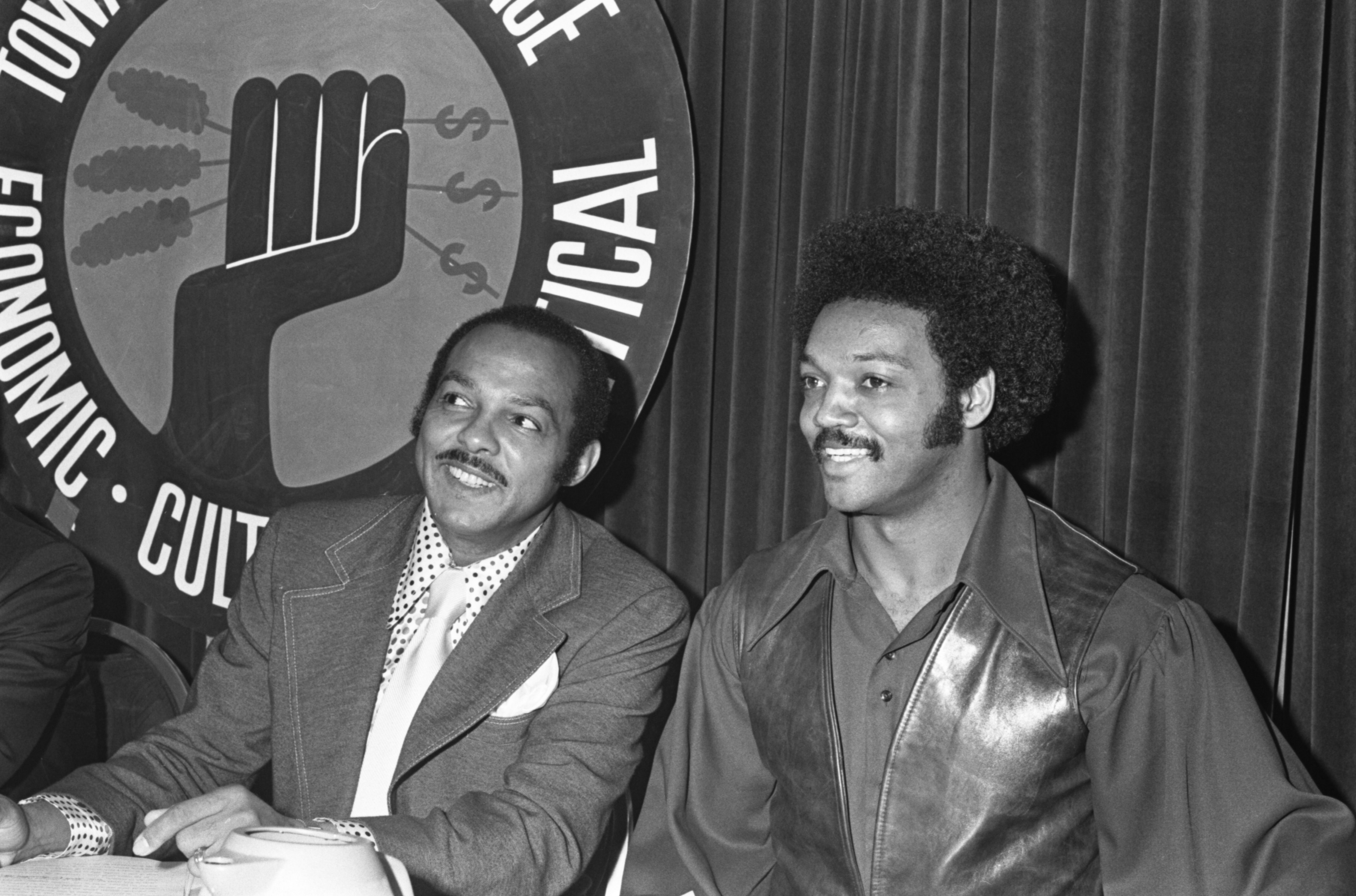 Two African American men in 1970s attire sit smiling at a table with a sign behind them showing a black fist with dollar signs and wheat, with words including "Economic" and "Political."