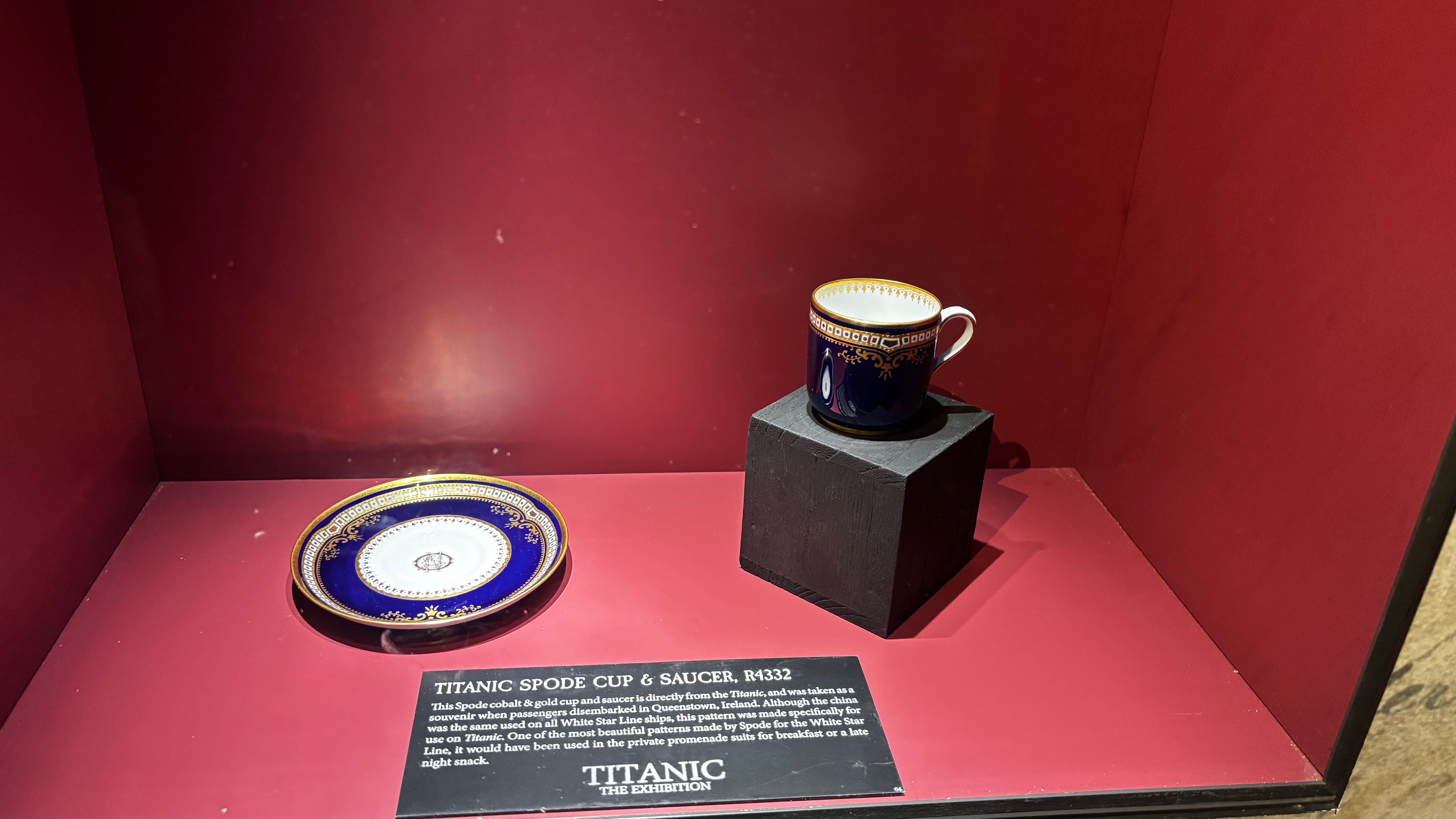 Cobalt blue and gold Spode cup and saucer from the Titanic, displayed on a red surface with an informational plaque in a museum exhibit titled "Titanic The Exhibition".