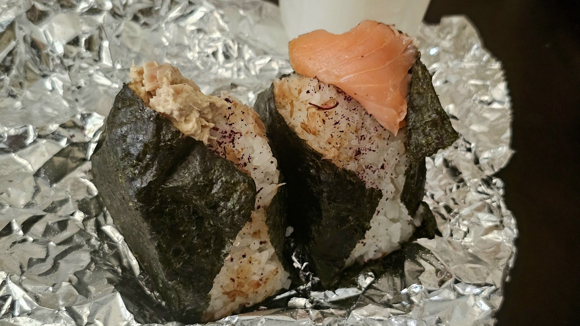 Two onigiri (rice balls) wrapped in nori, one topped with a slice of salmon; rice speckled with furikake, set on crinkled aluminum foil.