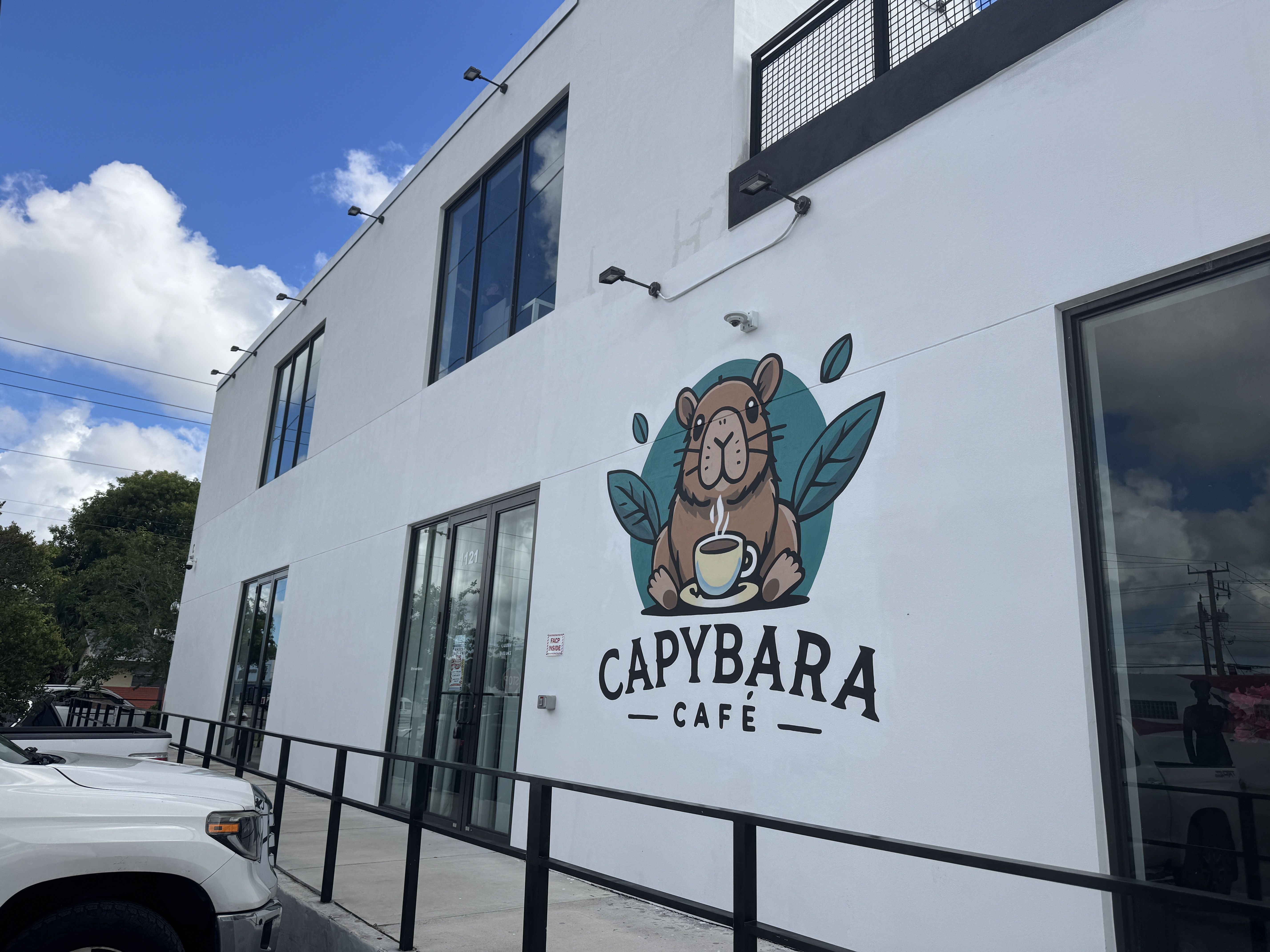 Bright white building with large glass windows and a mural of a brown capybara sipping coffee, surrounded by teal leaves; a sign on the wall reads "CAPYBARA CAFÉ" under a clear blue sky.
