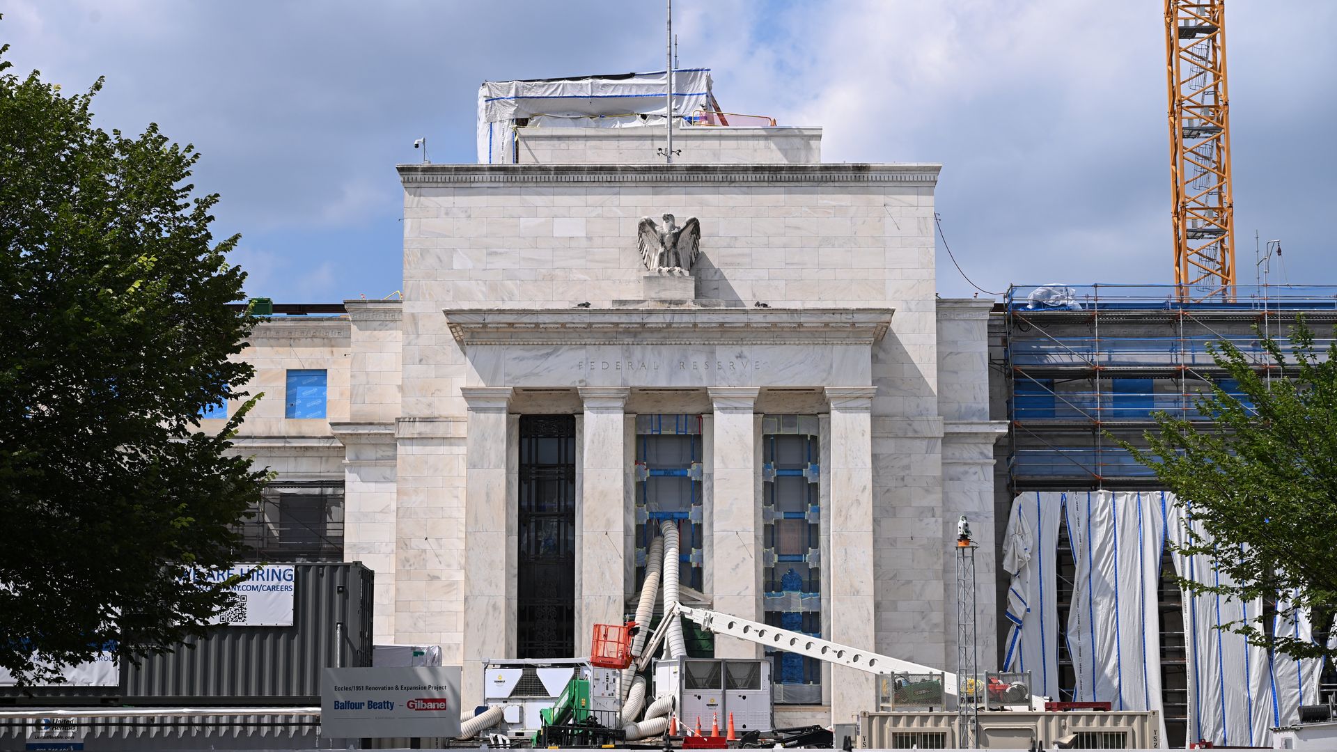 Renovation work on a large white marble Federal Reserve building with an American flag on top, construction equipment, scaffolding, and blue sky with clouds in the background.