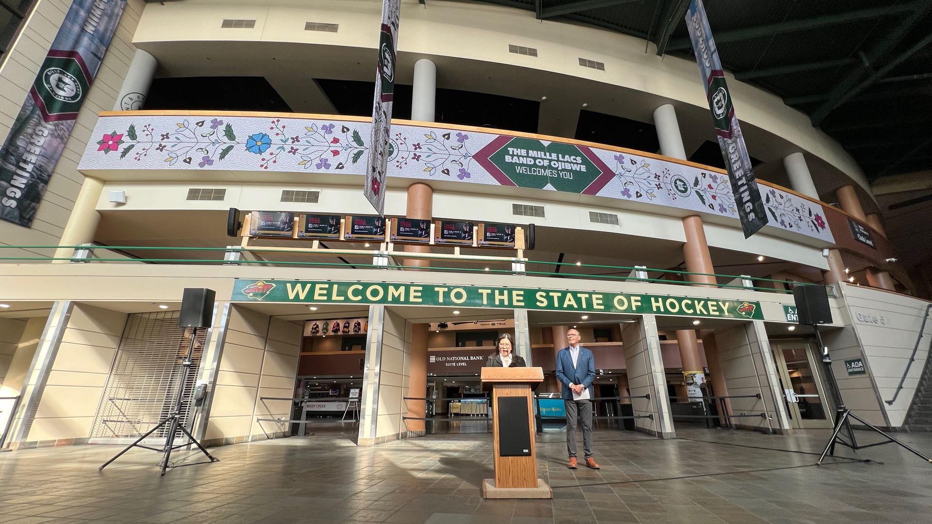 Inside a large arena lobby, two people stand at a podium beneath a green banner reading "WELCOME TO THE STATE OF HOCKEY." A floral balcony banner arches above with banners and speakers nearby.