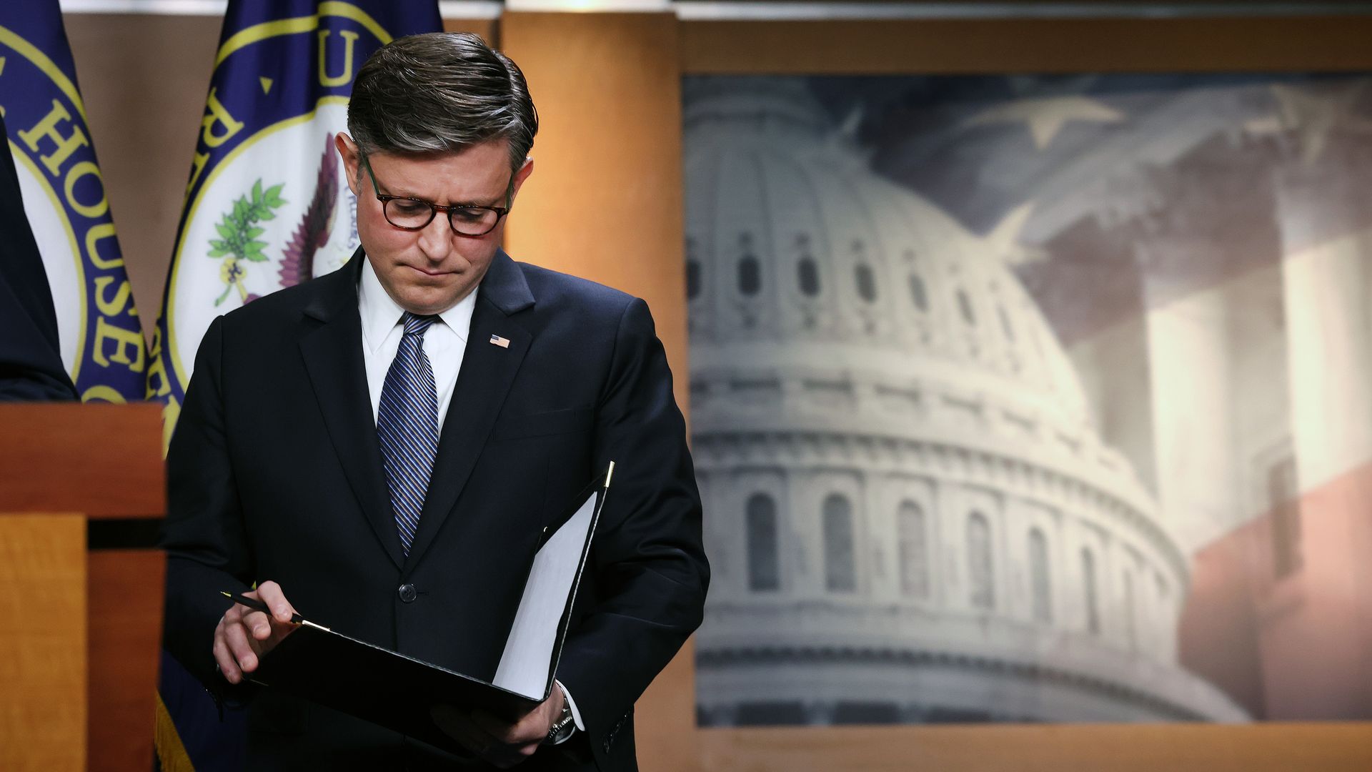 Man in a dark suit and glasses signs papers on a clipboard at a podium. Behind him are blue-and-gold flags and a mural of the Capitol dome.