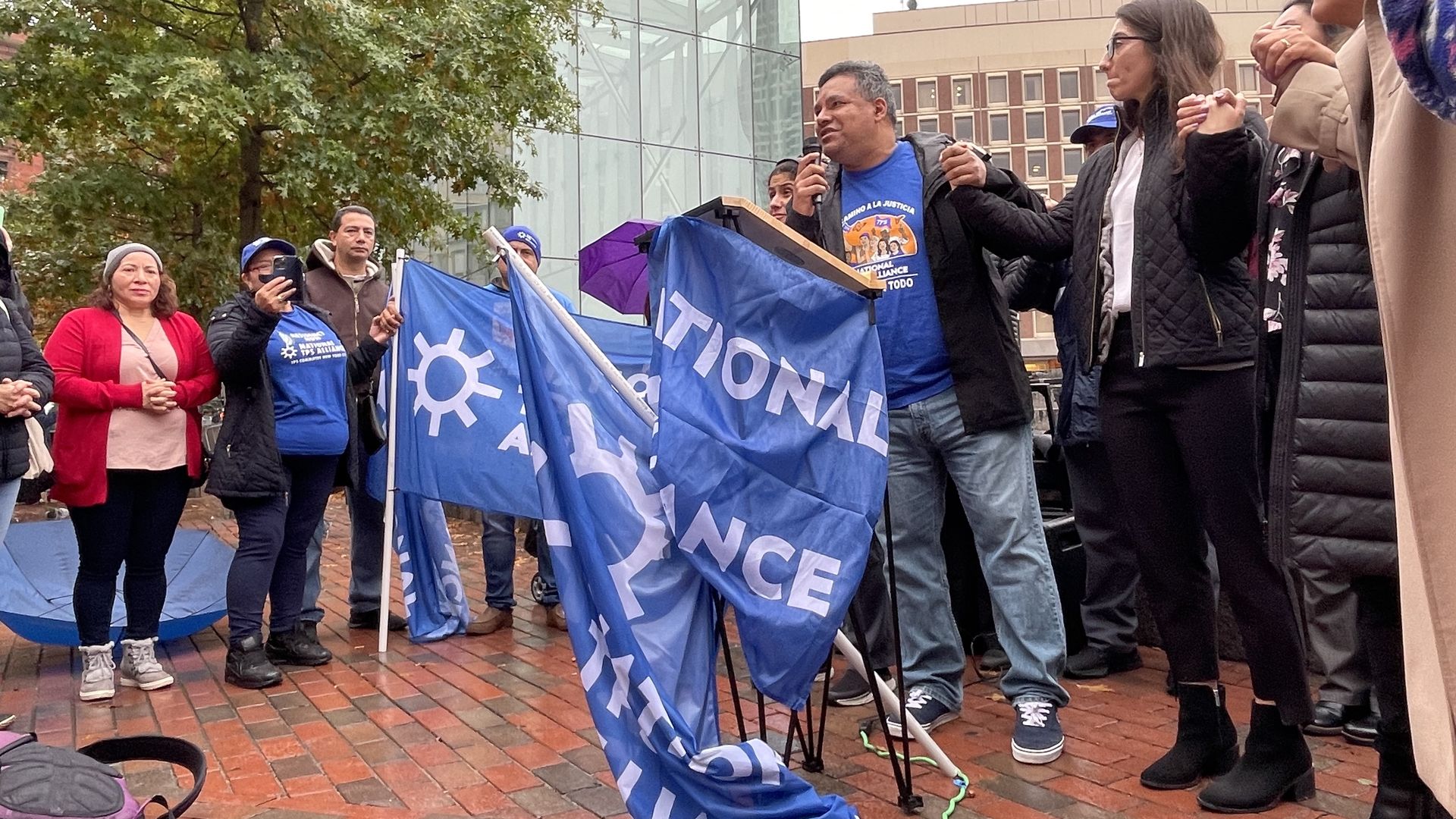 TPS holder Jose Palma, center, holds hands with Boston City Councilor Gabriela Coletta as he stands behind a podium.