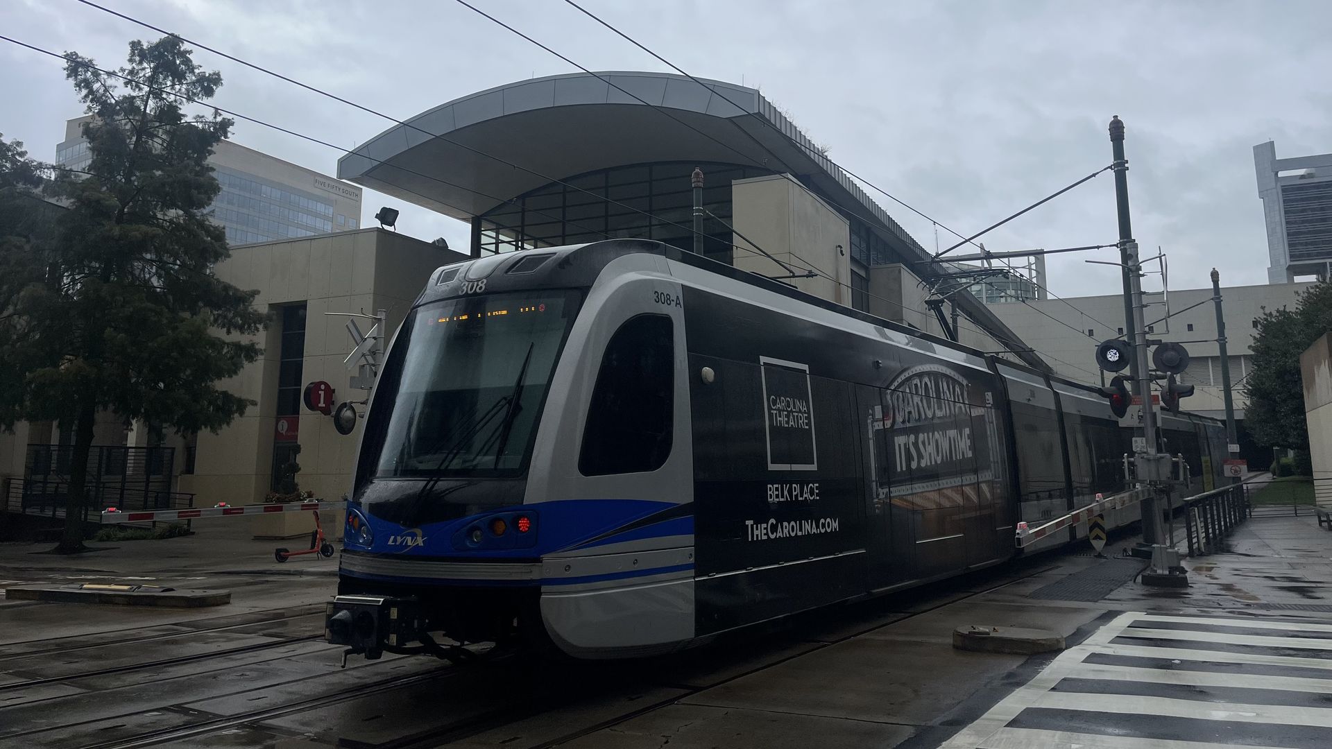 Blue, black, and white LYNX light rail train stopped at a wet urban street crossing with buildings and cloudy sky in the background.