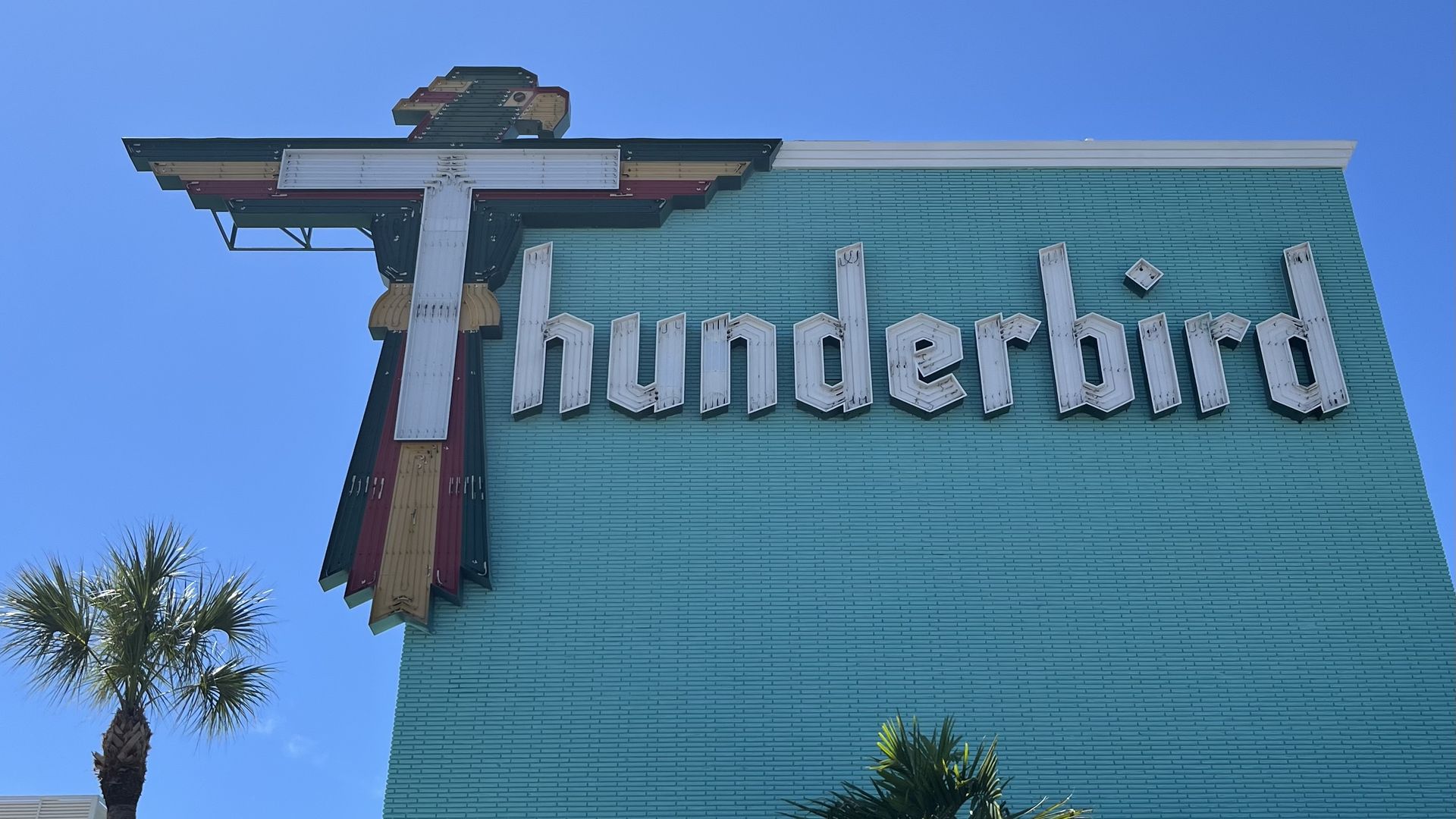 A teal brick wall with a large neon sign that says "Thunderbird." The T is decorated to look like a bird with red, green and yellow feathers.