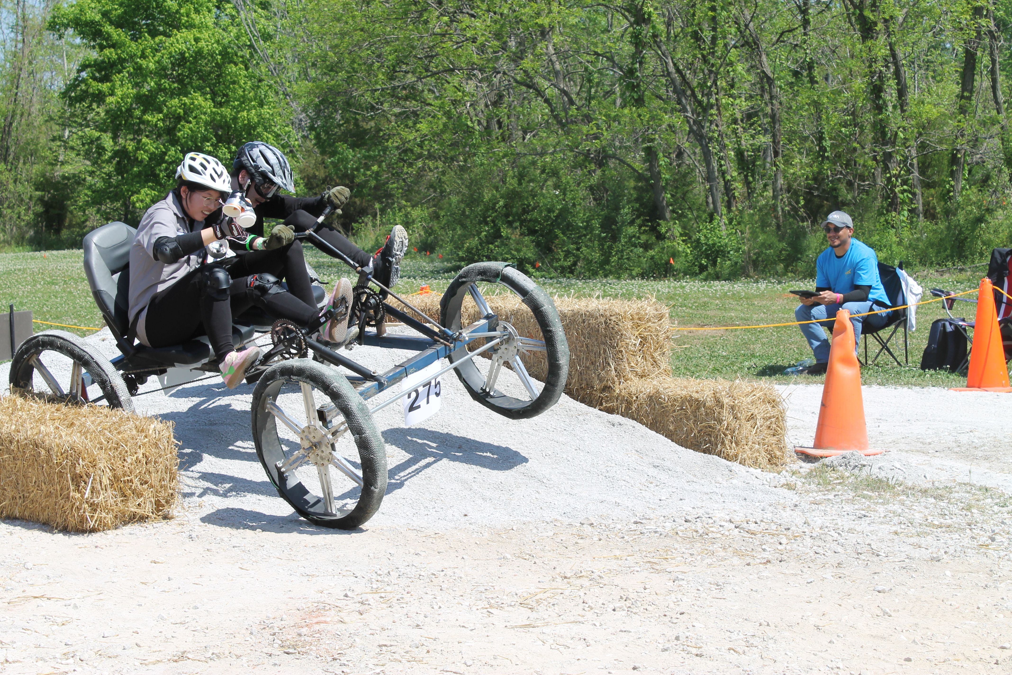 Two riders wearing helmets on a student-built, human-powered rover vehicle navigate a hay-bale obstacle course on gravel, as a man in a blue shirt sits with a clipboard and orange cones mark the course.