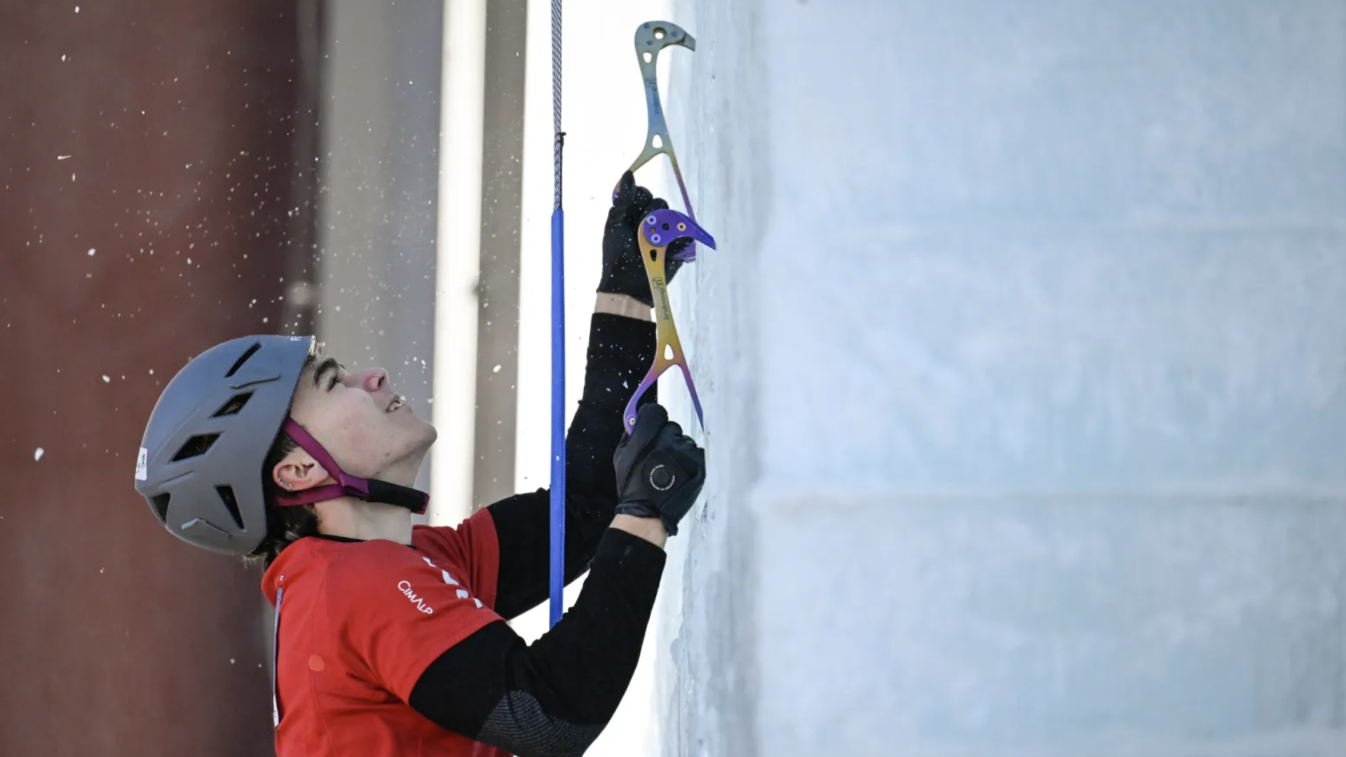 Person in a red shirt and gray helmet ice climbing on a frozen wall using ice tools, with snow particles in the air against a blurred background.