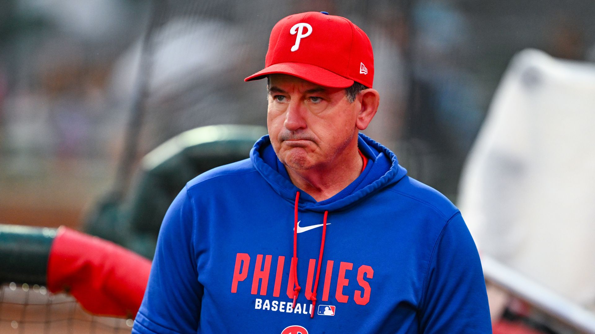 A man wearing a red Phillies baseball cap and a blue Phillies hoodie stands outdoors with a serious expression; blurred background shows a fence and dugout.