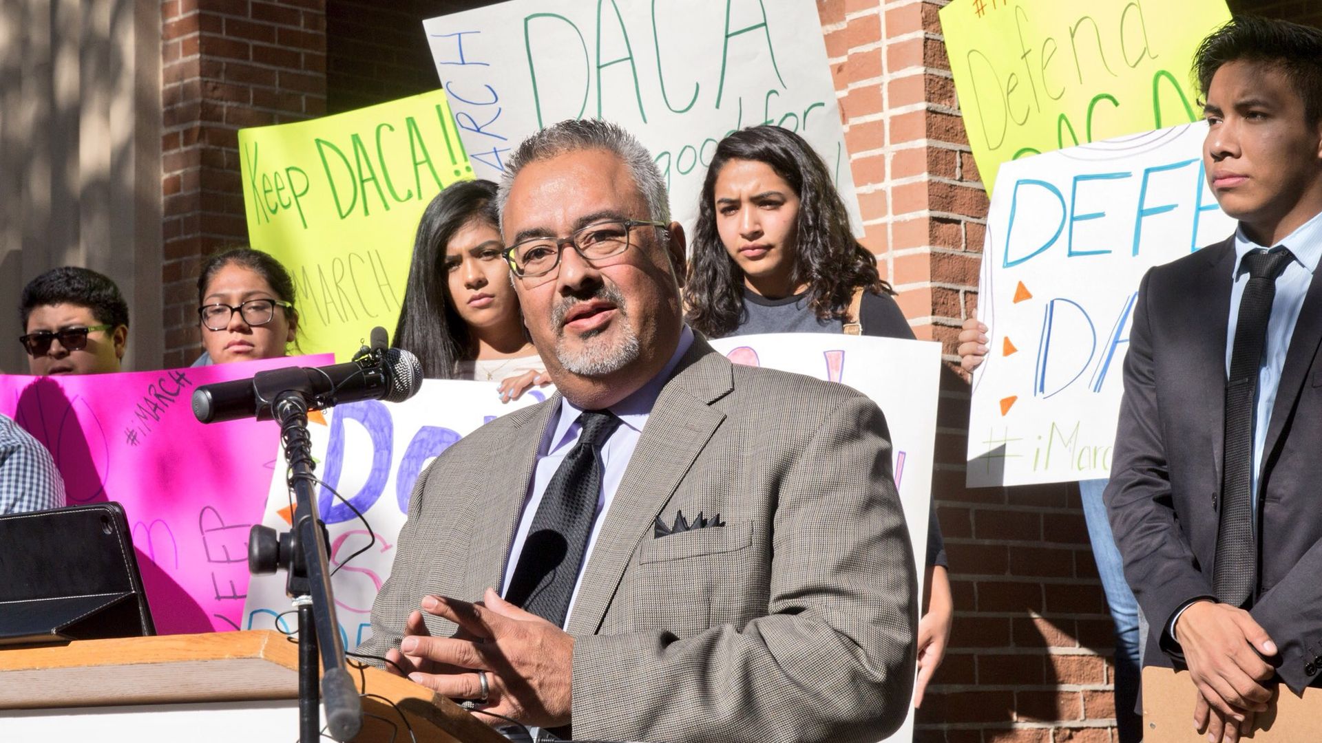 Man in a gray suit speaking at a podium with a microphone, surrounded by young people holding signs supporting DACA, including messages like "Keep DACA" and "Defend DACA."