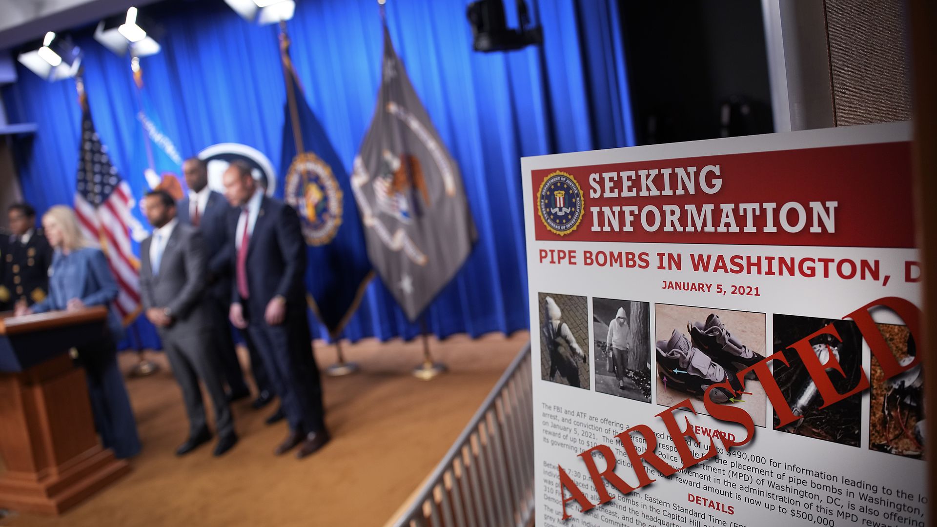 A poster with information on the Jan. 5, 2021 pipe bomber reads "ARRESTED" in red font as government officials are seen in the background during a news conference. 