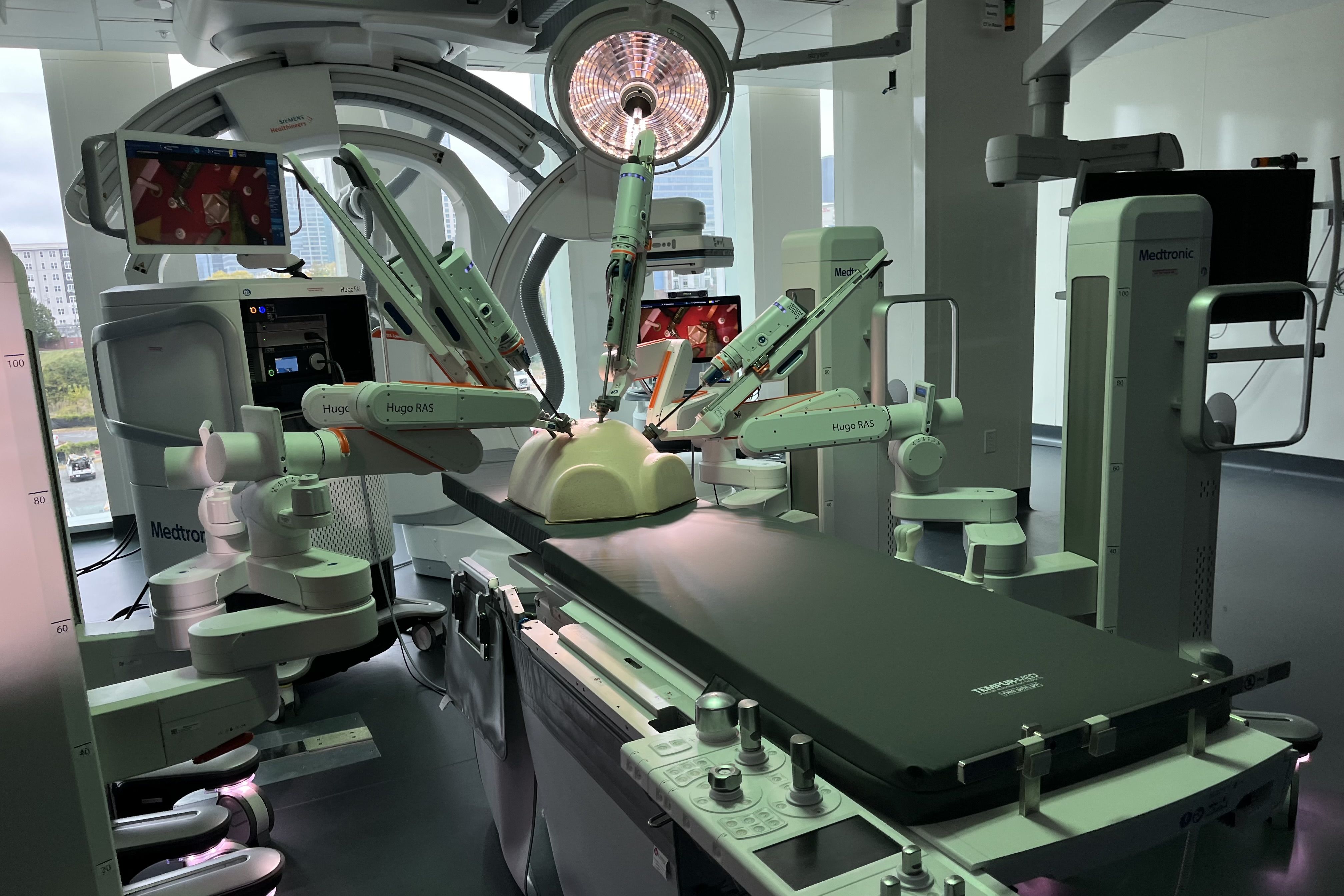 Modern operating room with a robotic surgical system labeled Hugo RAS by Medtronic, robotic arms positioned over a padded surgical table under bright surgical lights, and monitors displaying images.