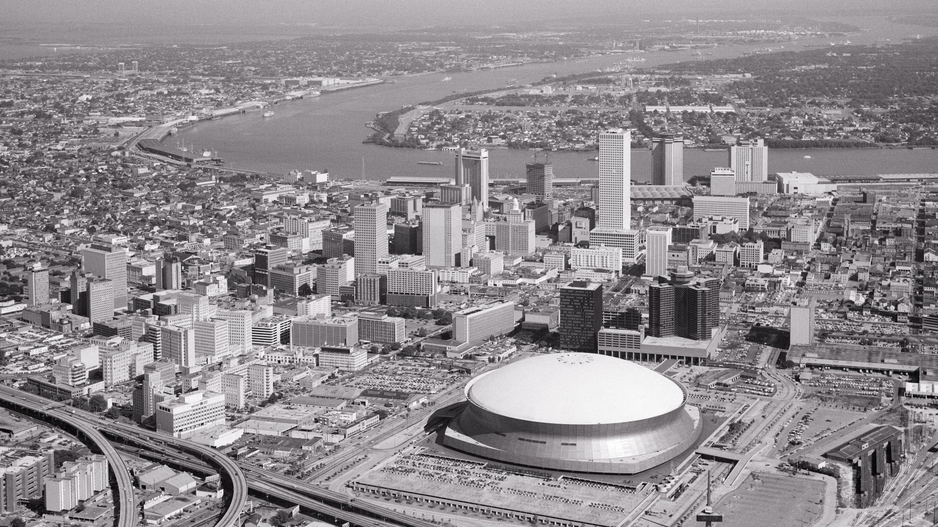 Black and white aerial view of New Orleans with a large domed stadium in the foreground, surrounded by highways, tall buildings, and a river in the background.