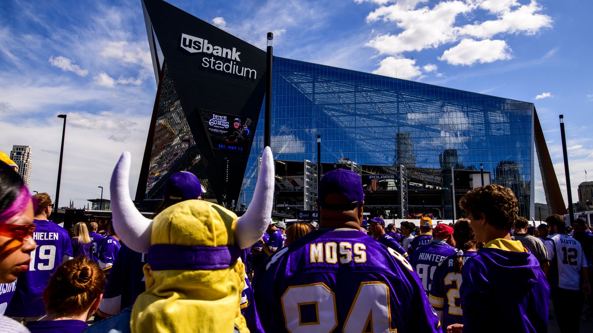 Crowd of Minnesota Vikings fans in purple jerseys outside US Bank Stadium, a modern blue-glass venue with a sharp black angular entrance, under a bright blue sky.