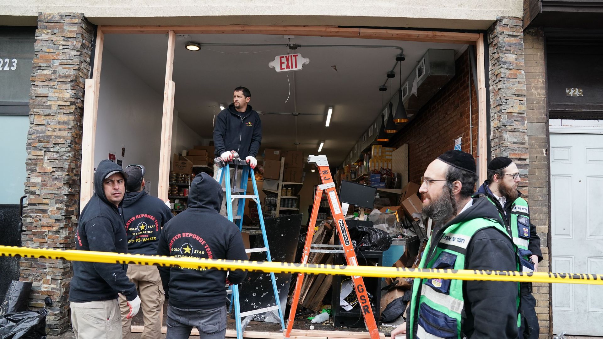 In this image, men wearing safety vests and hoodies stand in front of a large, open storefront window and one man stands on a ladder