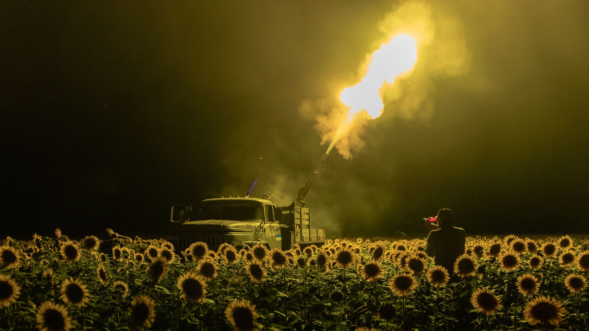 Nighttime scene of a military truck firing a large gun in a field of sunflowers, with a person in the foreground photographing the scene under yellow-orange light from the gunfire.