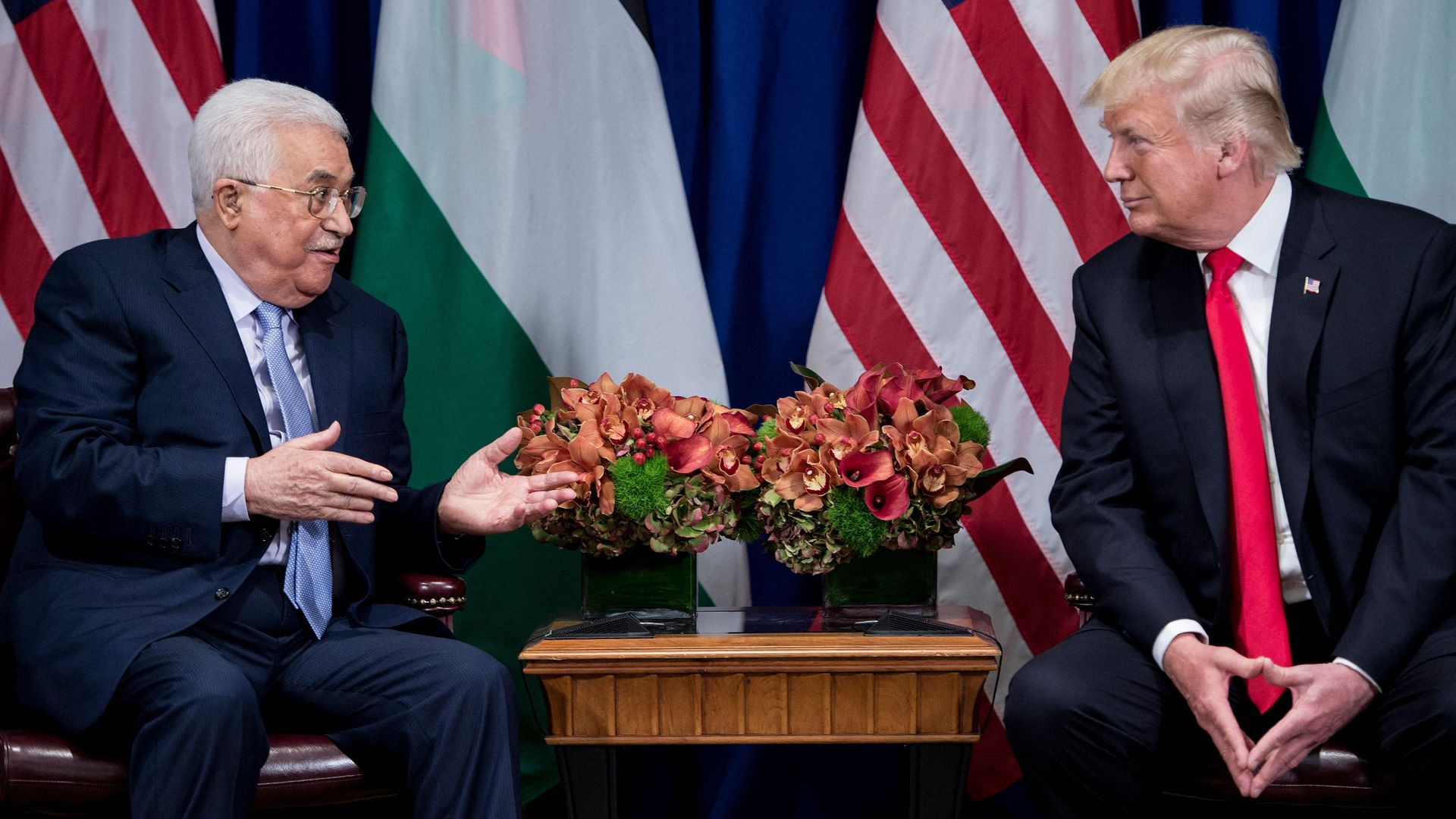 Two men in dark suits sit facing each other with a small wooden table and floral arrangements between them, with U.S. and Palestine flags behind them.