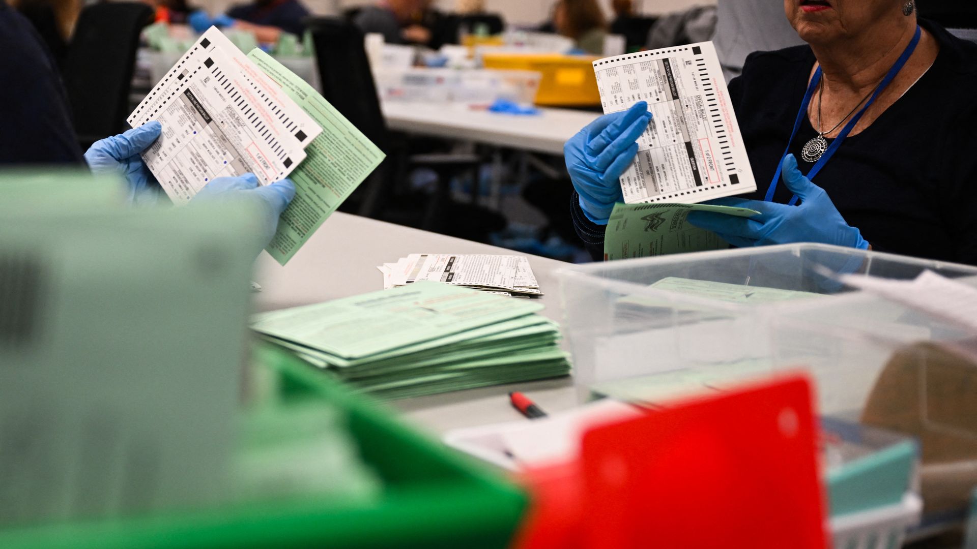 Election workers wearing blue gloves sort and examine ballots and green envelopes that contain them at a table with multiple bins on it. 