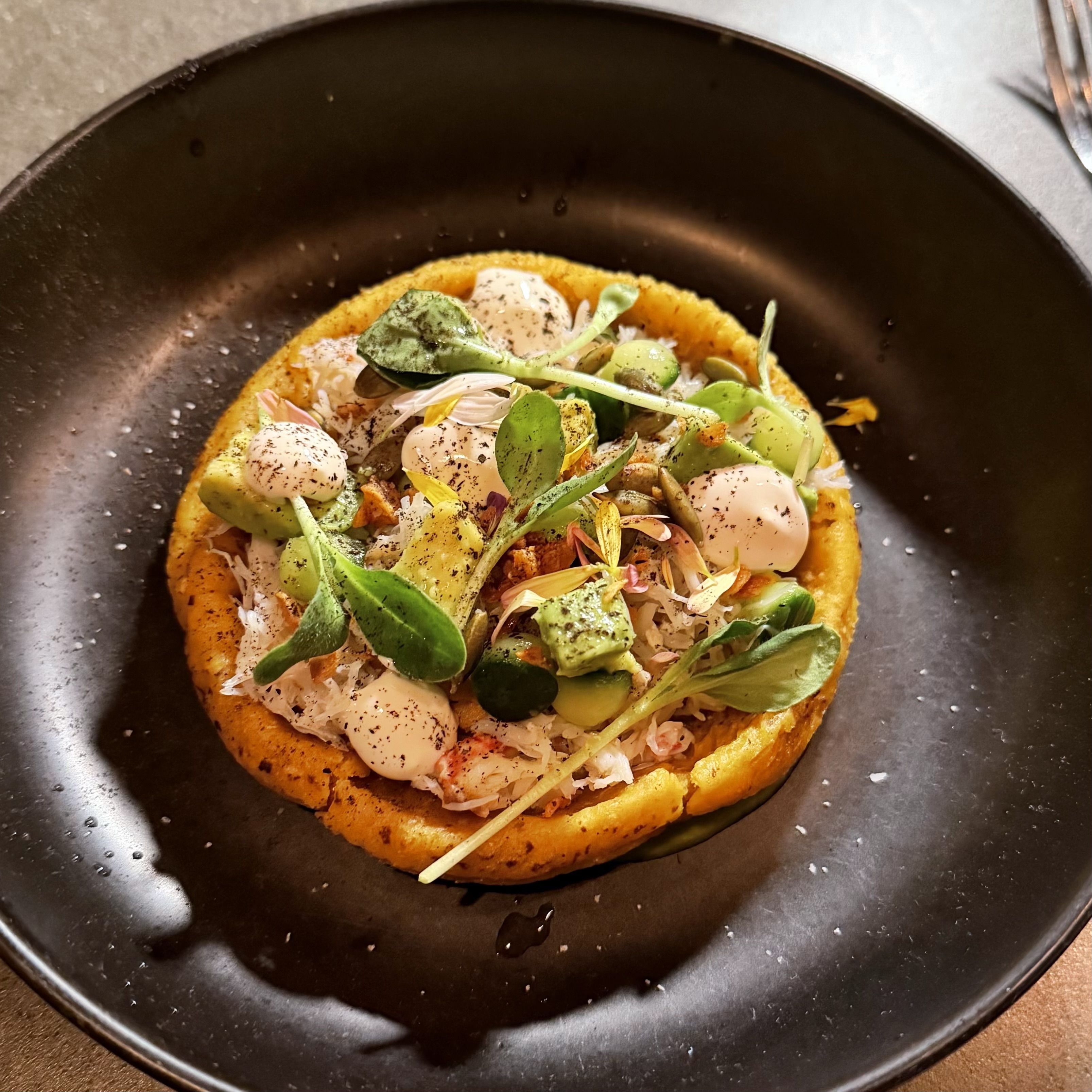 Gourmet dish on a black plate featuring a crab base topped with green leafy herbs, white sauce dollops, and sprinkled black pepper, with an orange circular base underneath.