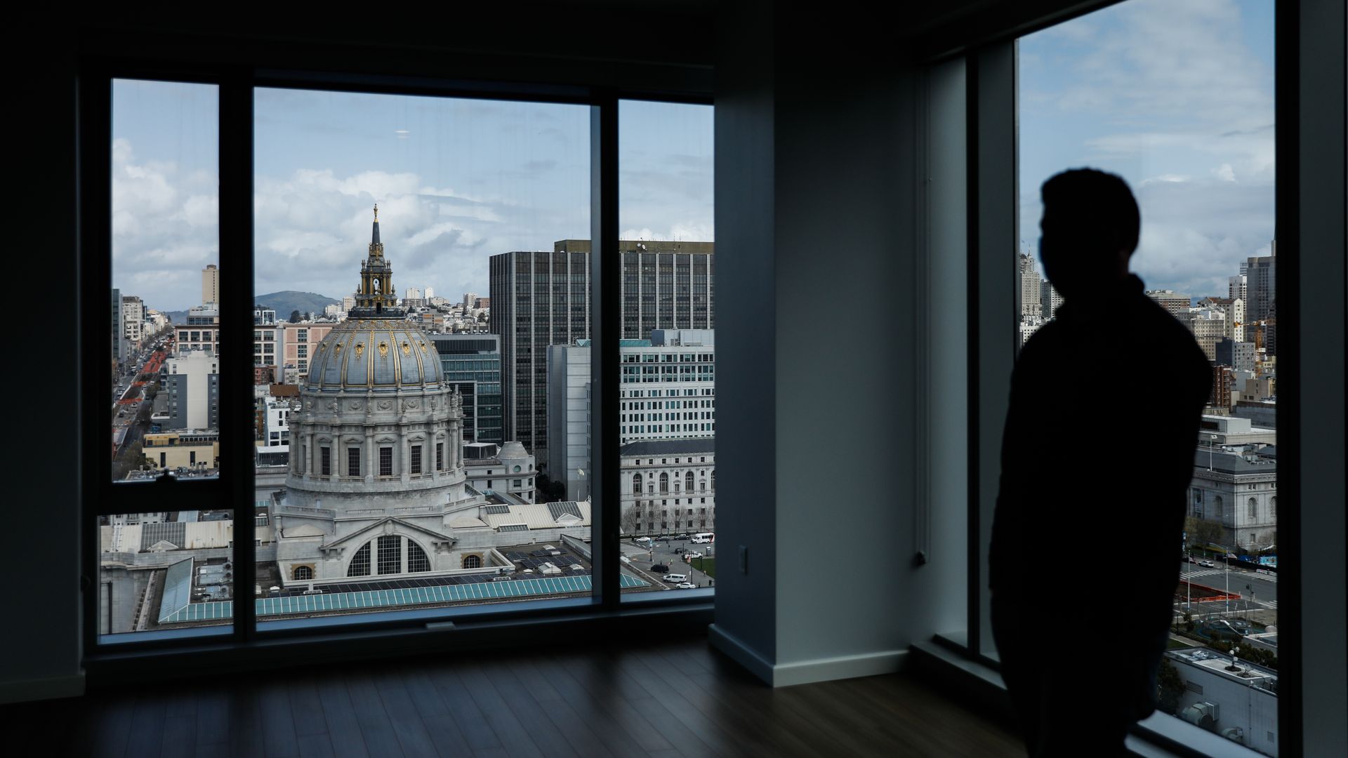 Photo of a silhouetted person standing in an empty room as they look out at the SF City Hall building